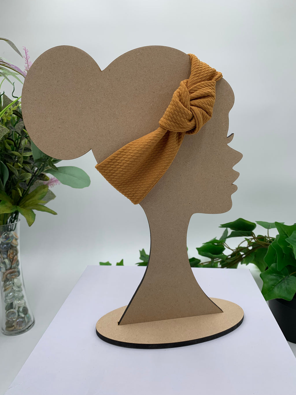 Mustard colored knot headband on a mannequin head silhouette against a white background with green leaves