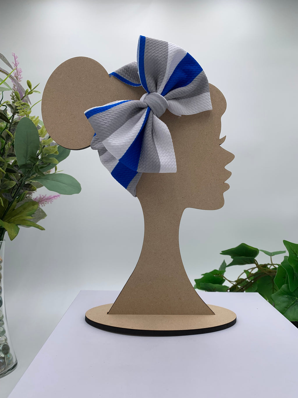Wooden mannequin head cutout with a blue, white, and gray bow, against a white background with greenery.