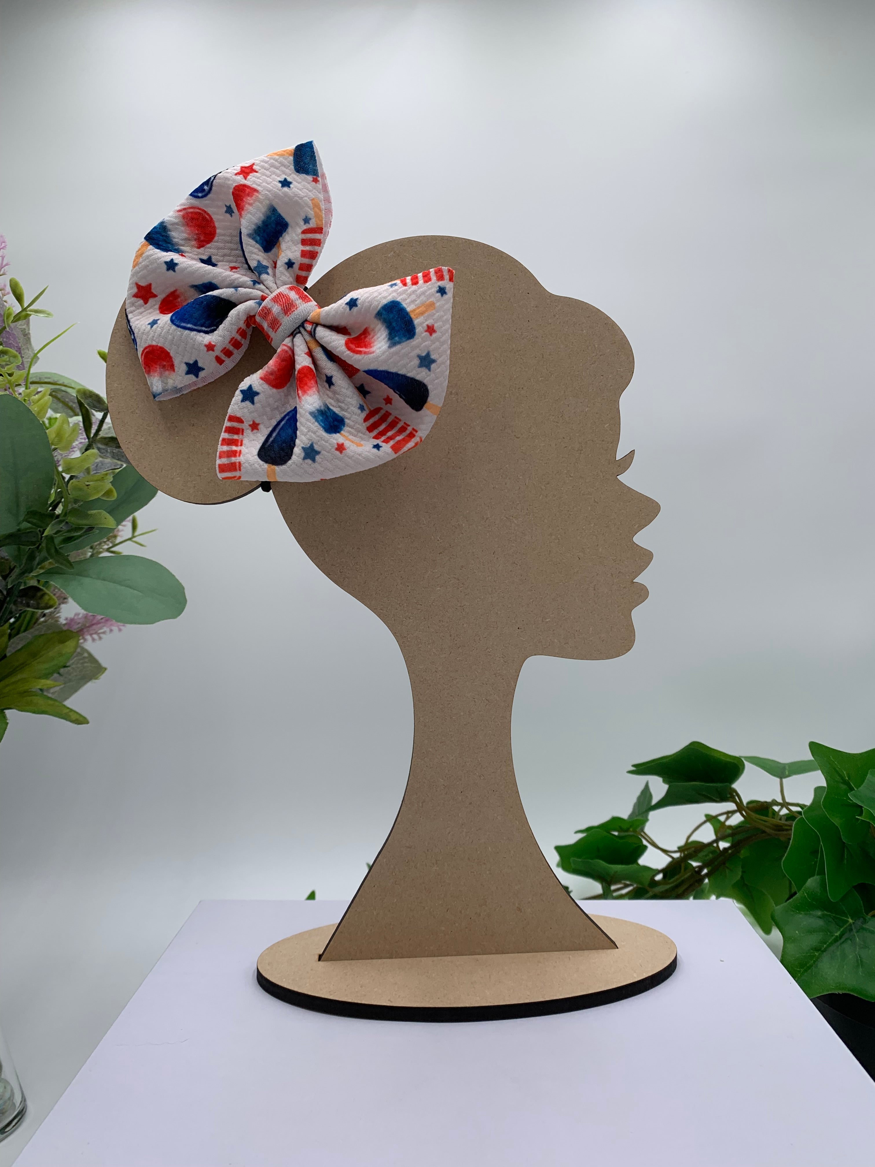 Multicolored 4th of july themed hair bow on a wooden silhouette of a head with plants in the background