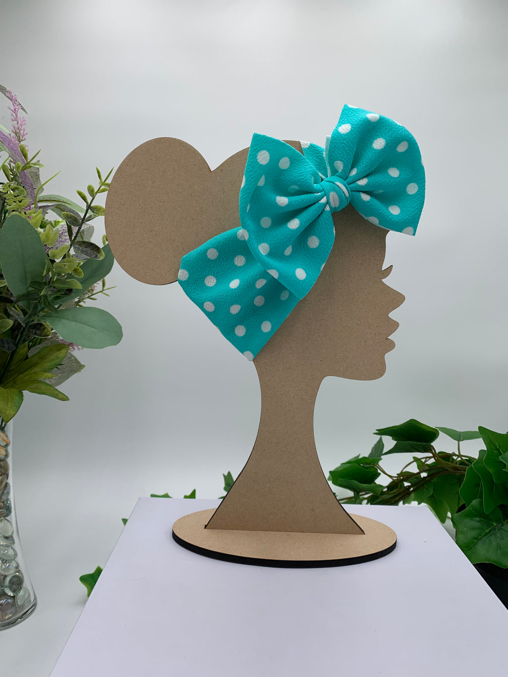 teal with white polka dots themed big bow headband on a mannequin head with a white background and greenery 