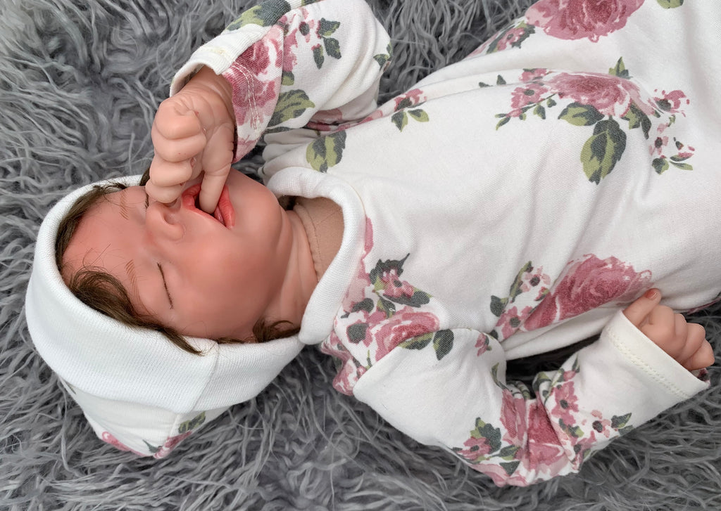 close up of Newborn baby doll in a Floral baby sleeper sack with matching beanie on a gray background with green leaves.