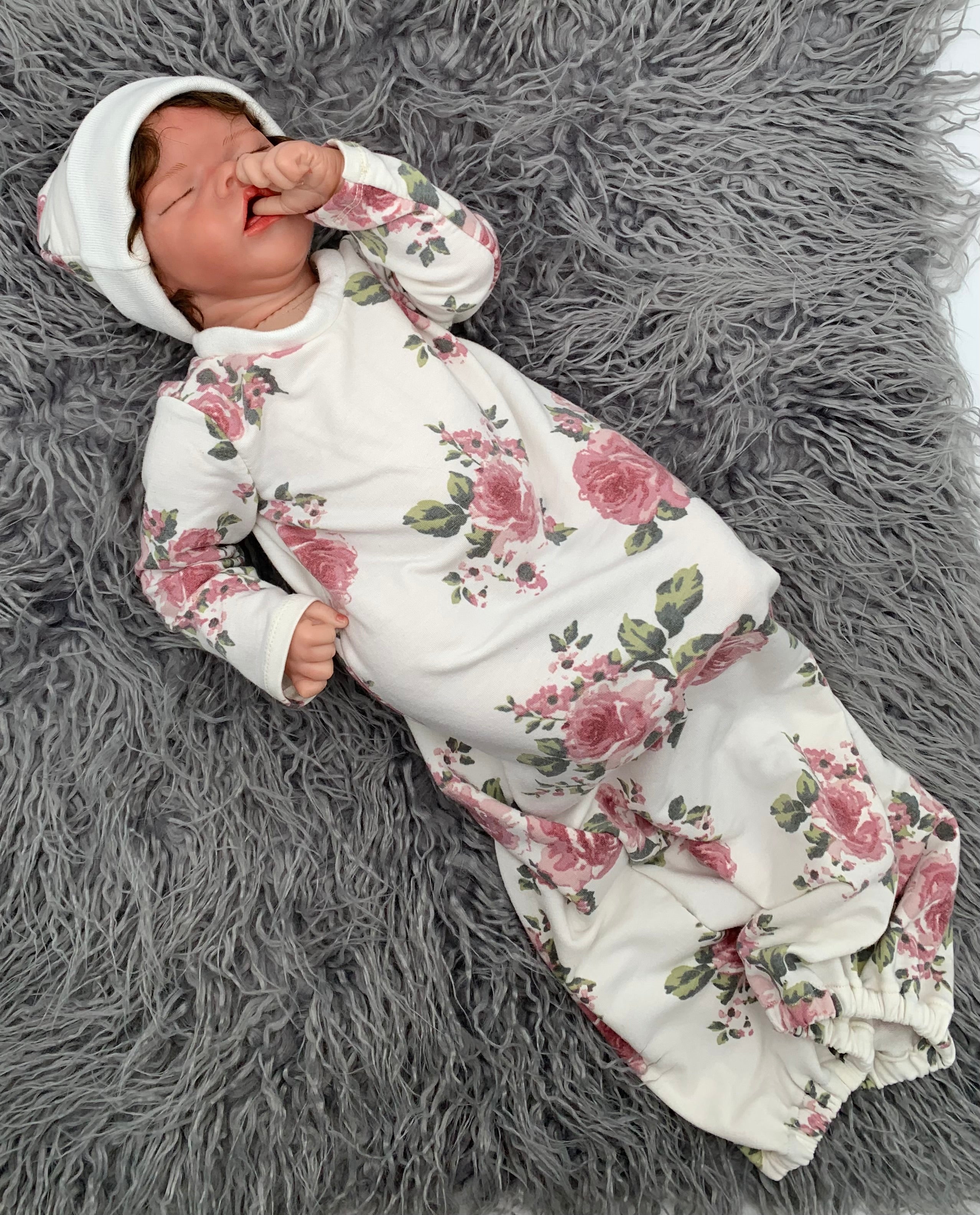 Newborn baby doll in a Floral baby sleeper sack with matching beanie on a gray background with green leaves.