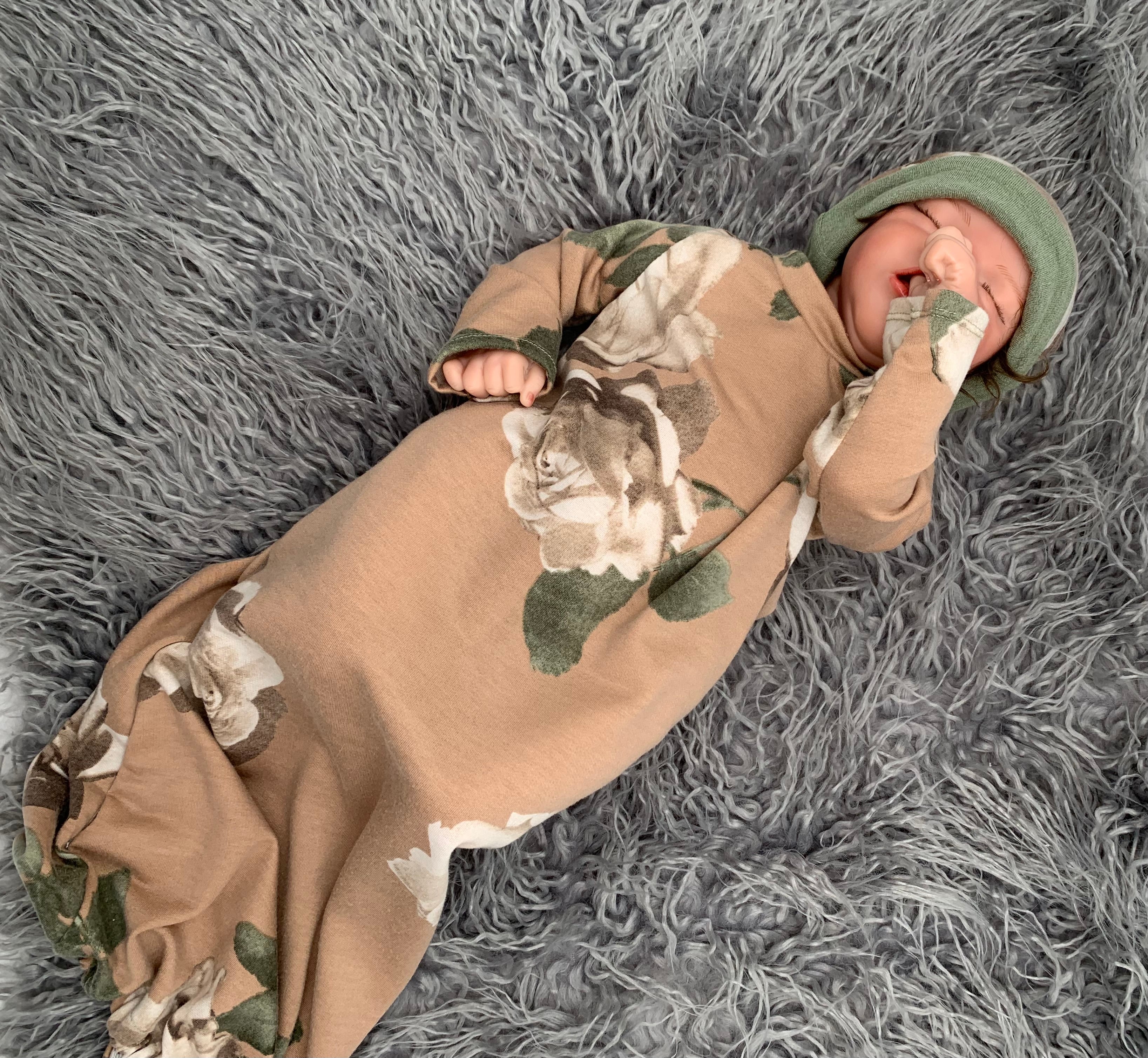 Newborn baby doll in a Floral baby sleeper sack with matching beanie on a gray background with green leaves.