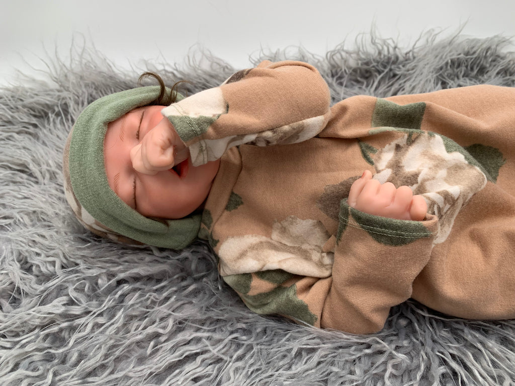 close up of a Newborn baby doll in a Floral baby sleeper sack with matching beanie on a gray background with green leaves.