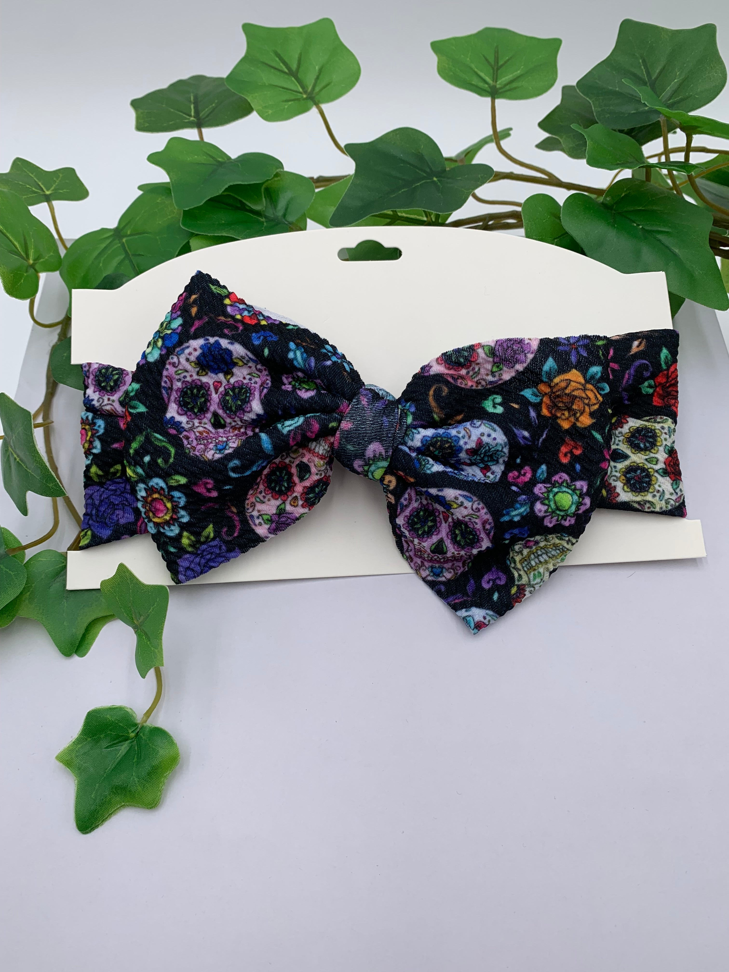 Day of the dead sugar skull themed big bow headband on a white card with a white background and greenery 