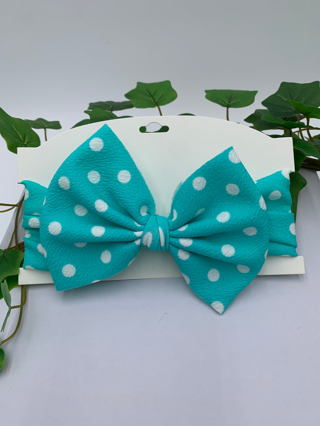teal with white polka dots themed big bow headband on a white card with a white background and greenery 