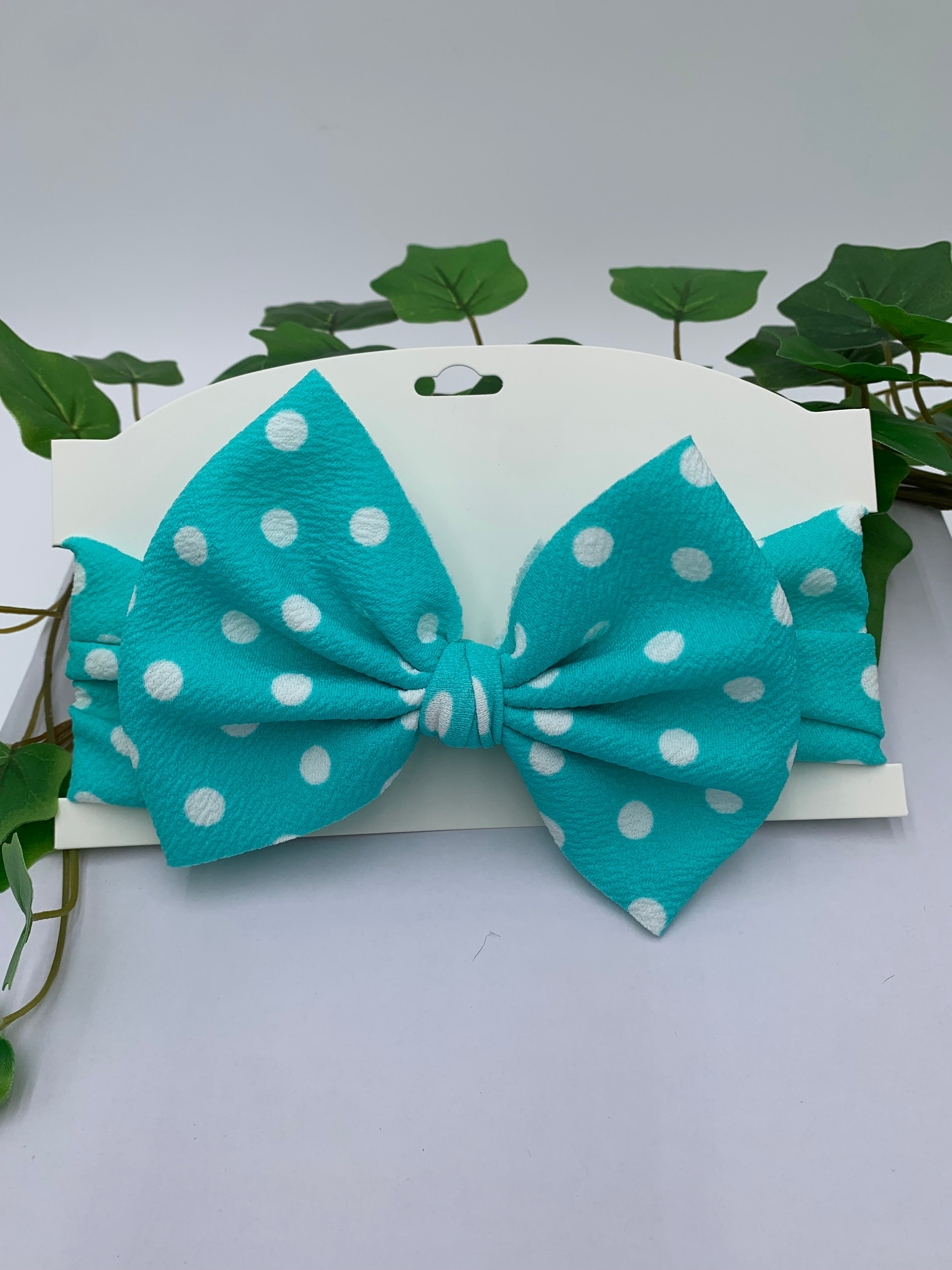 teal with white polka dots themed big bow headband on a white card with a white background and greenery 