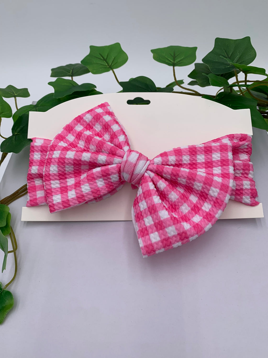 Pink plaid themed big bow headband on a white card with a white background and greenery 