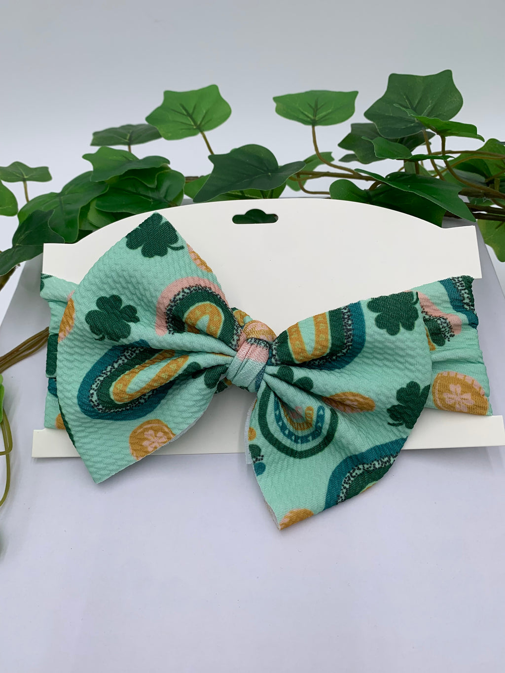 Green St. Patrick's Day themed big bow headband on a white card with a white background and greenery 