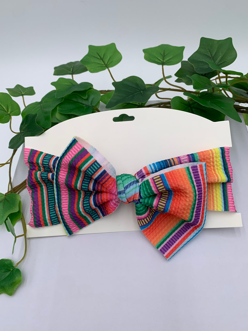 Colorful Serape themed big bow headband on a white card with a white background and greenery 