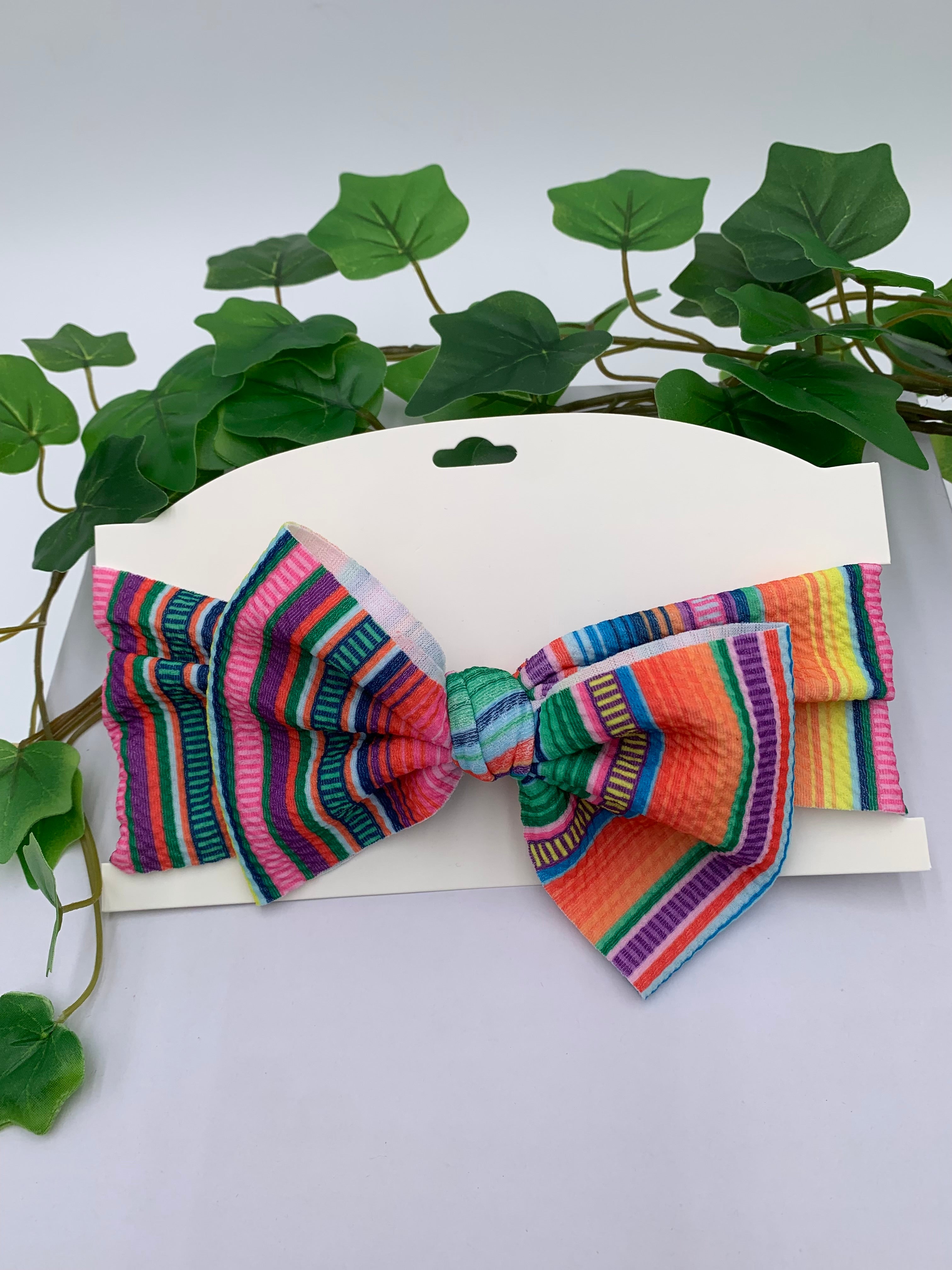 Colorful Serape themed big bow headband on a white card with a white background and greenery 