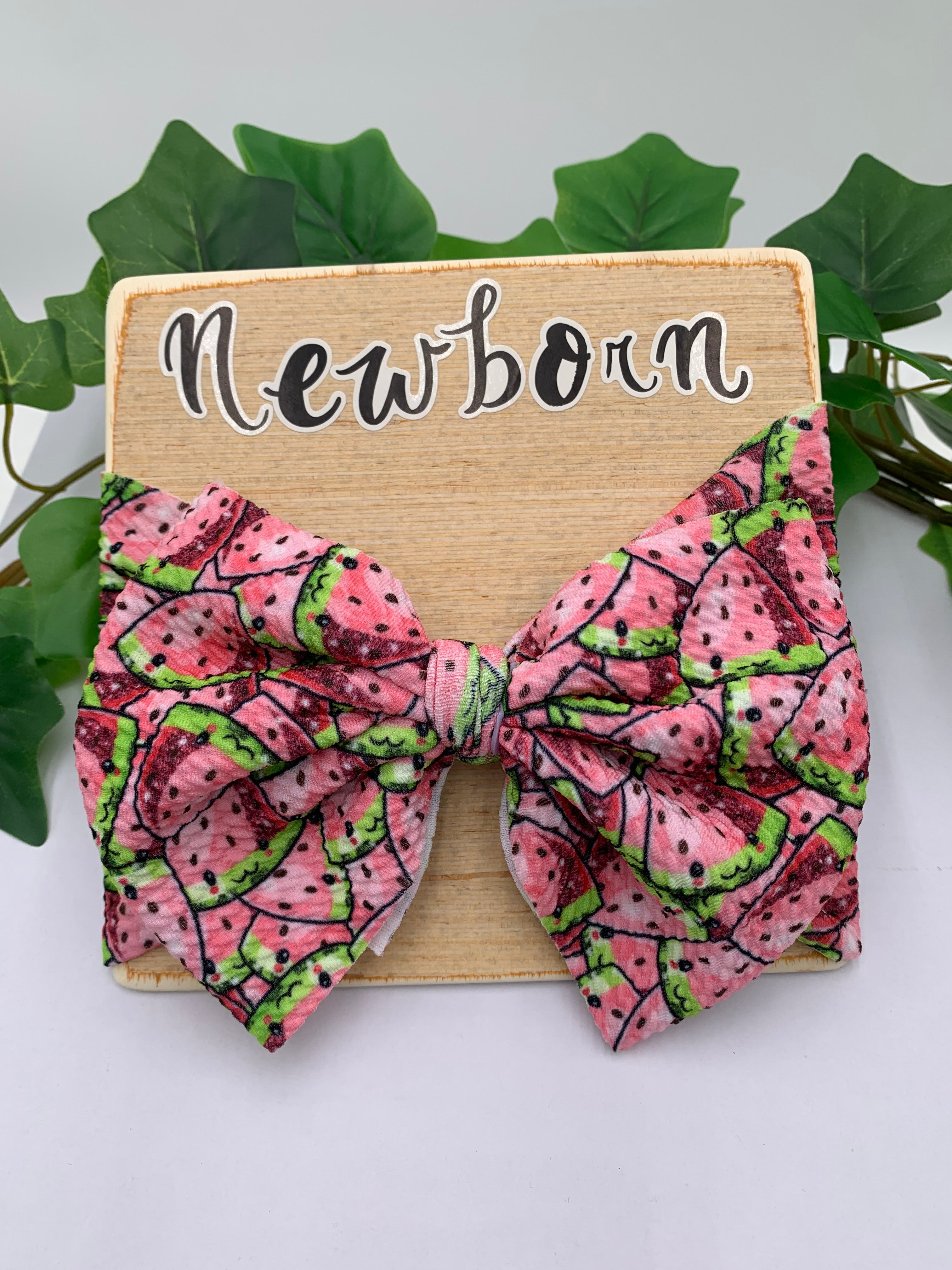 Watermelon slices themed big bow headband on a wooden board with text "newborn" with a white background and greenery 