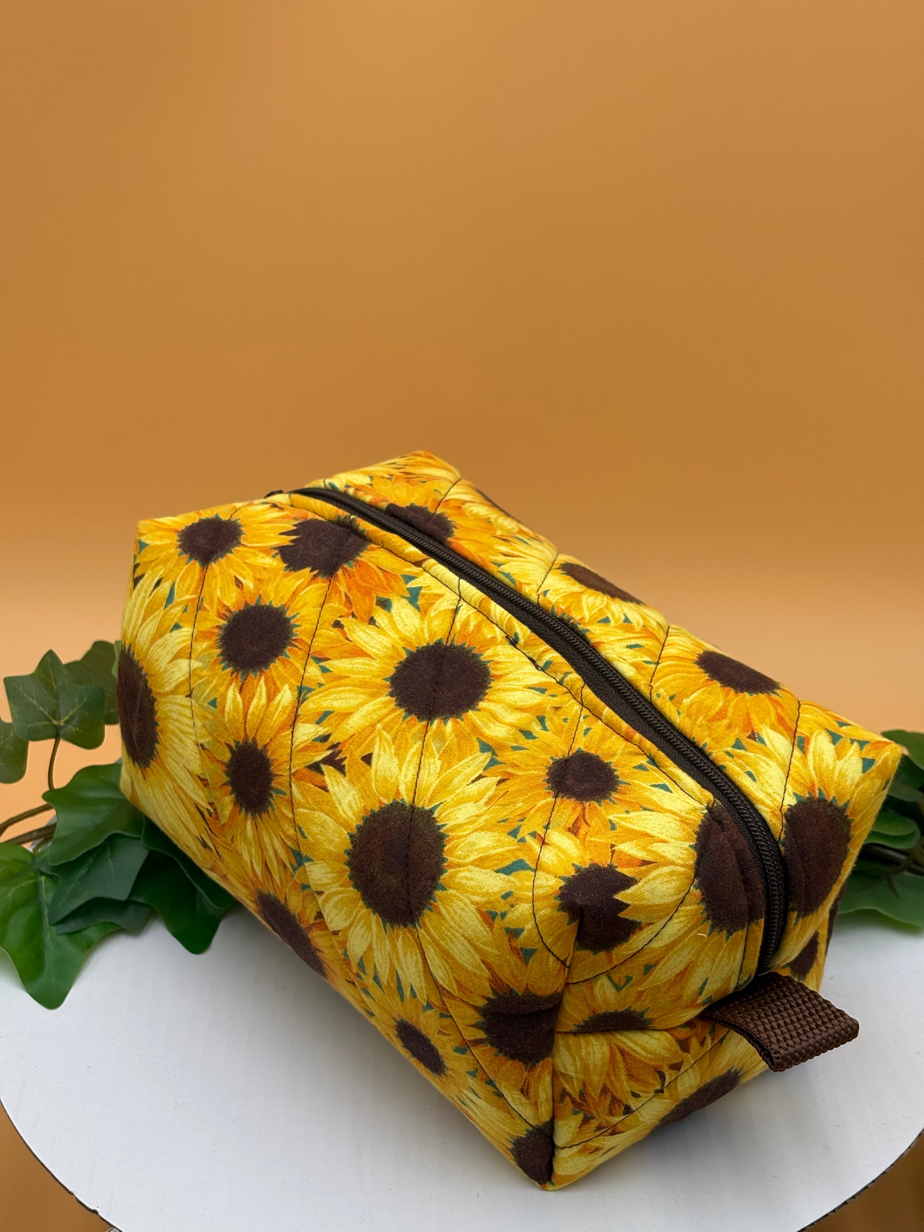 Quilted Sunflower pattern boxy makeup bag on a tan background with a potted green plant