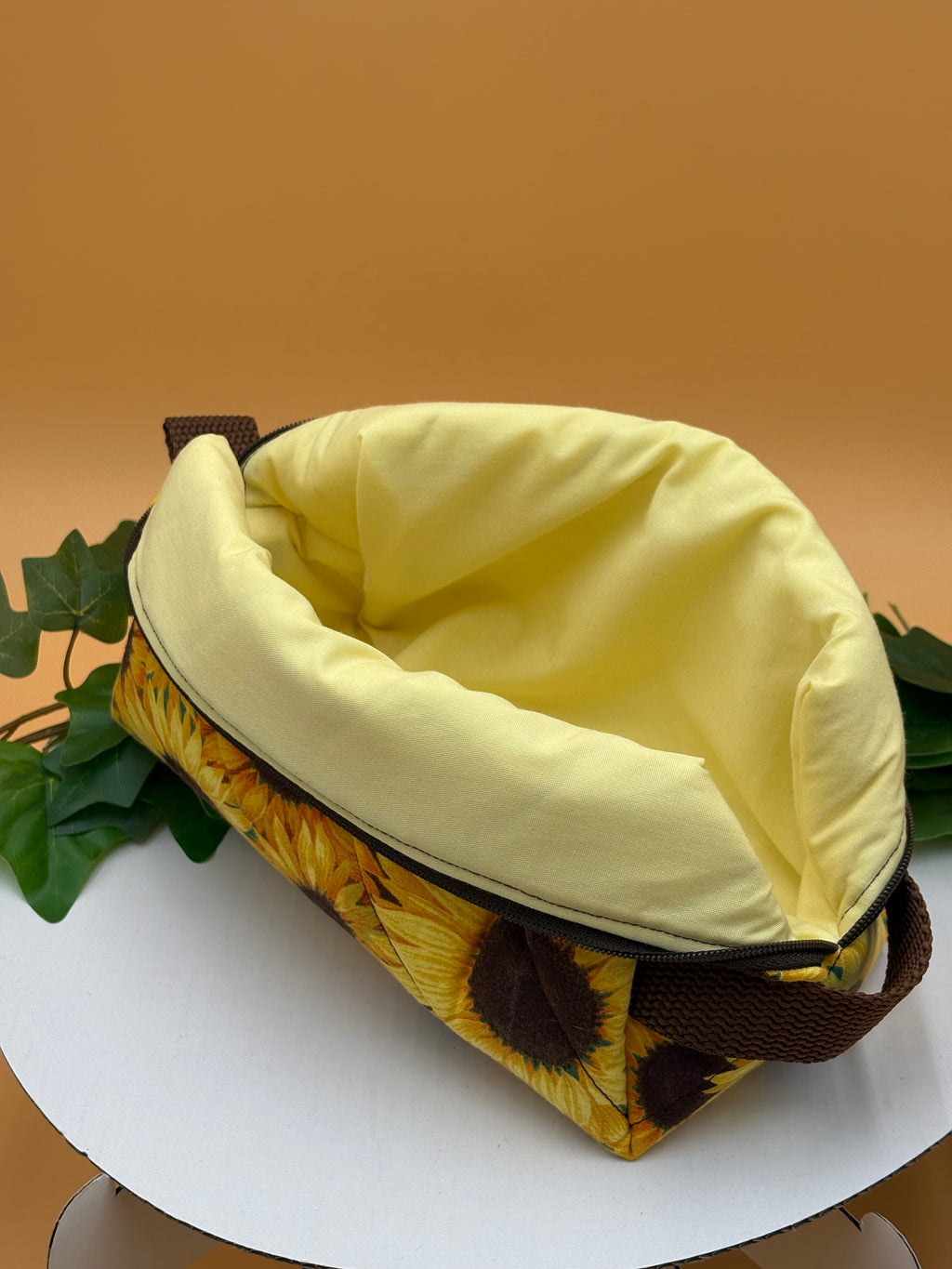 Quilted Sunflower pattern boxy makeup bag with pale yellow lining exposed on a tan background with a potted green plant