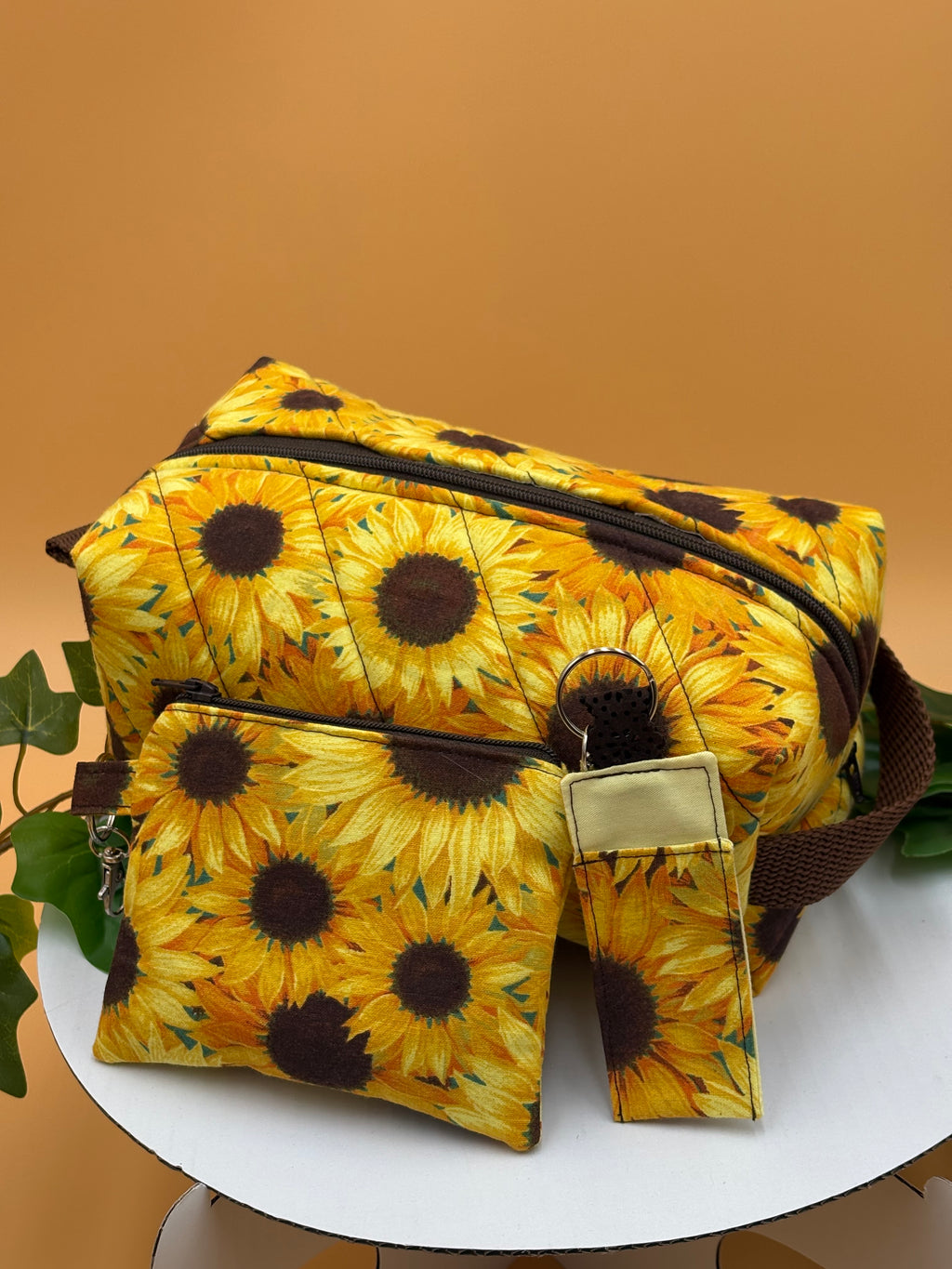 Quilted Sunflower pattern boxy makeup bag, coin purse keychain, and chapstick holder keychain with a on a tan background with a potted green plant
