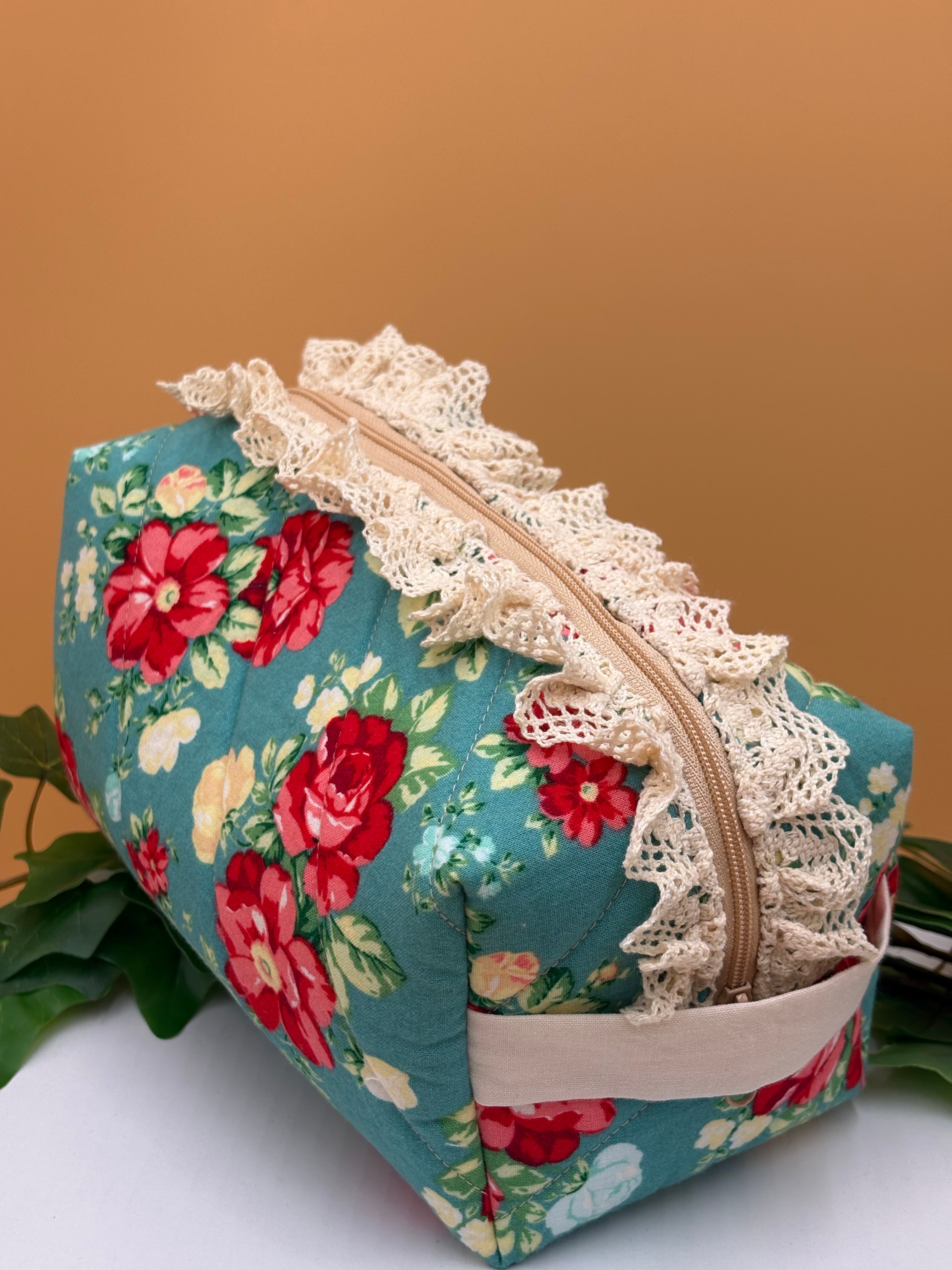Teal Floral fabric boxy makeup bag with lace trim on a tan background with green leaves