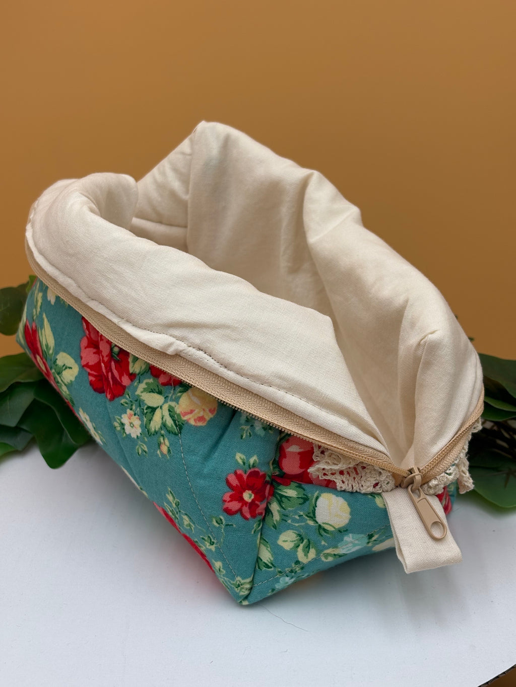Teal Floral fabric boxy makeup bag with a beige lining exposed on a tan background with green leaves