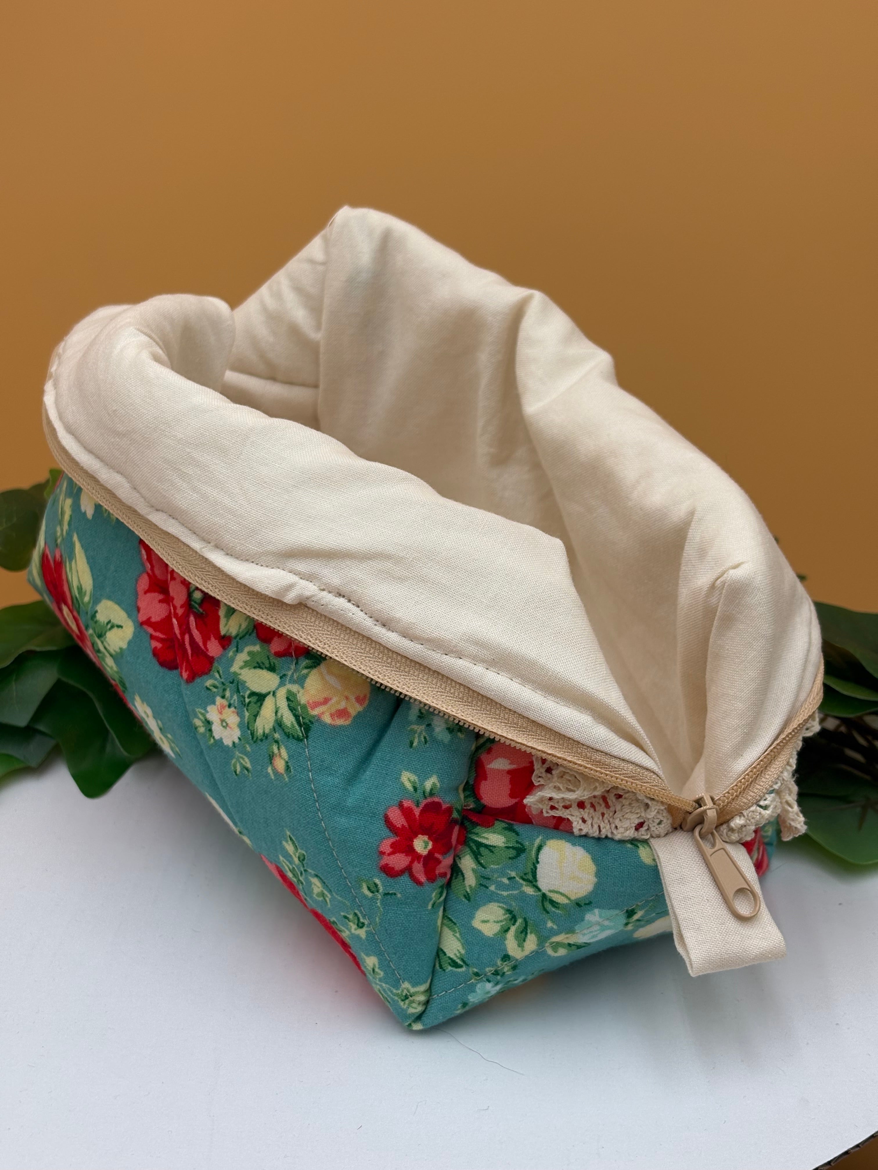 Teal Floral fabric boxy makeup bag with a beige lining exposed on a tan background with green leaves
