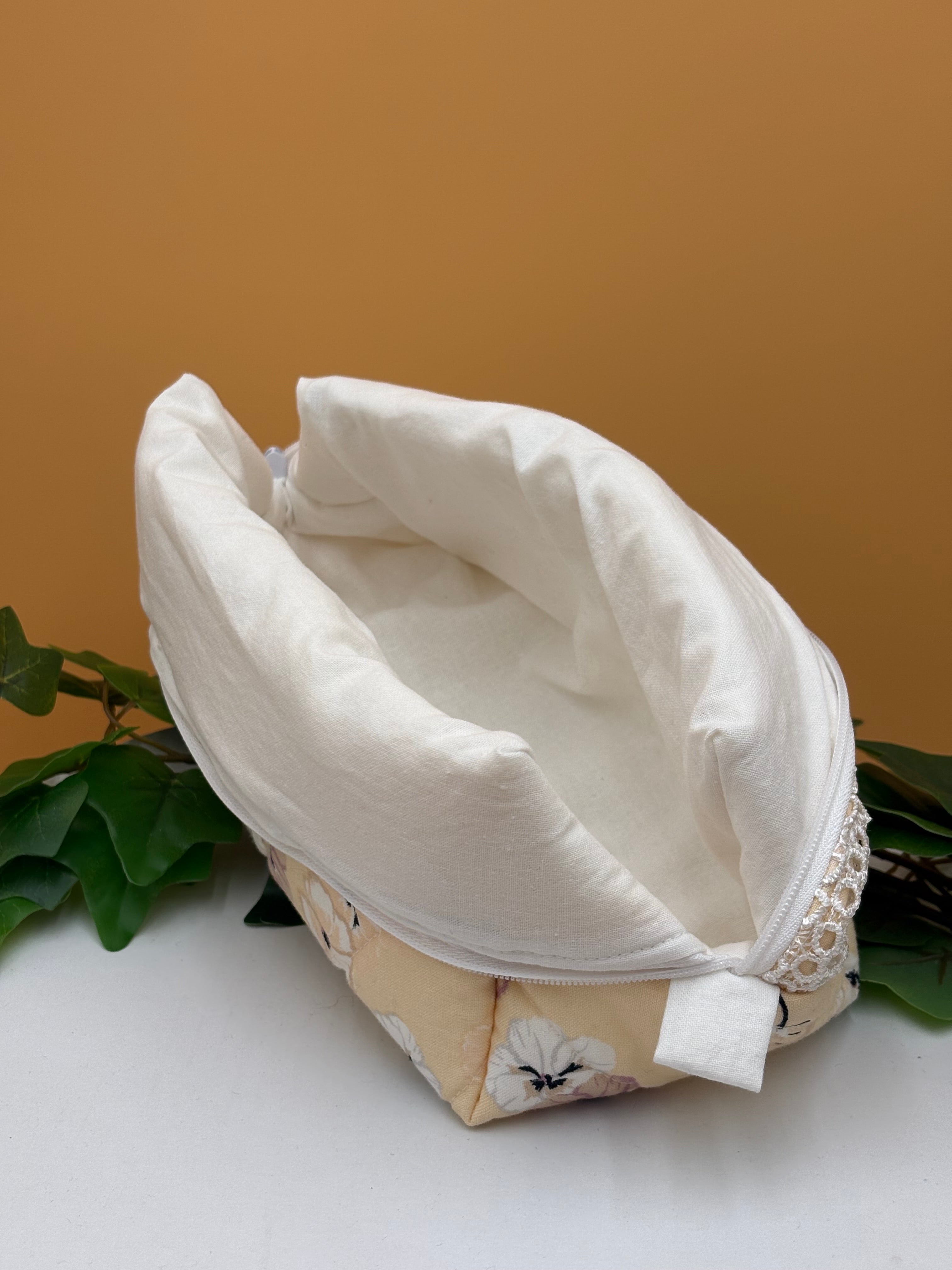 Pale Peach Floral fabric boxy makeup bag with a white lining exposed on a tan background with green leaves