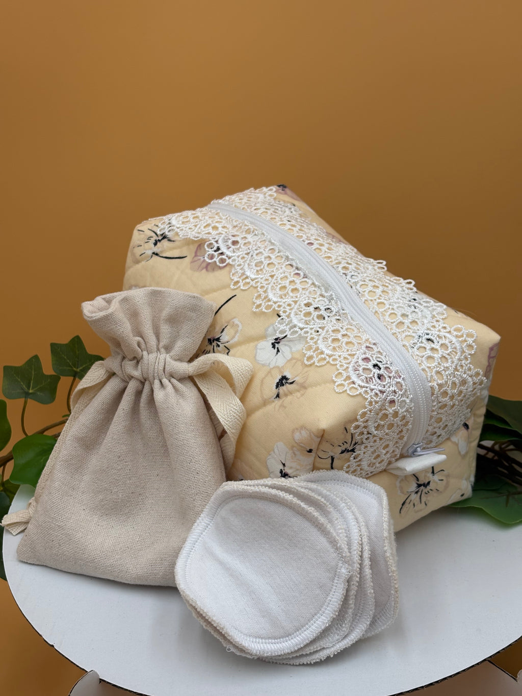 Pale Peach Floral fabric boxy makeup bag with a white lace, beige drawstring bag and 5 facial rounds on a tan background with green leaves