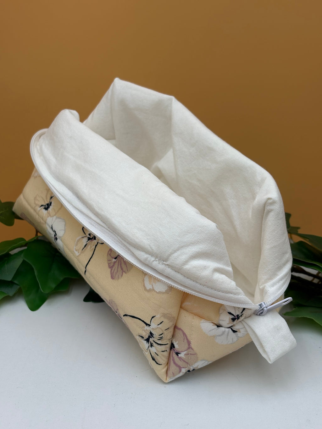 Quilted Pale Peach Floral pattern boxy makeup bag with white lining exposed on a tan background with a potted green plant