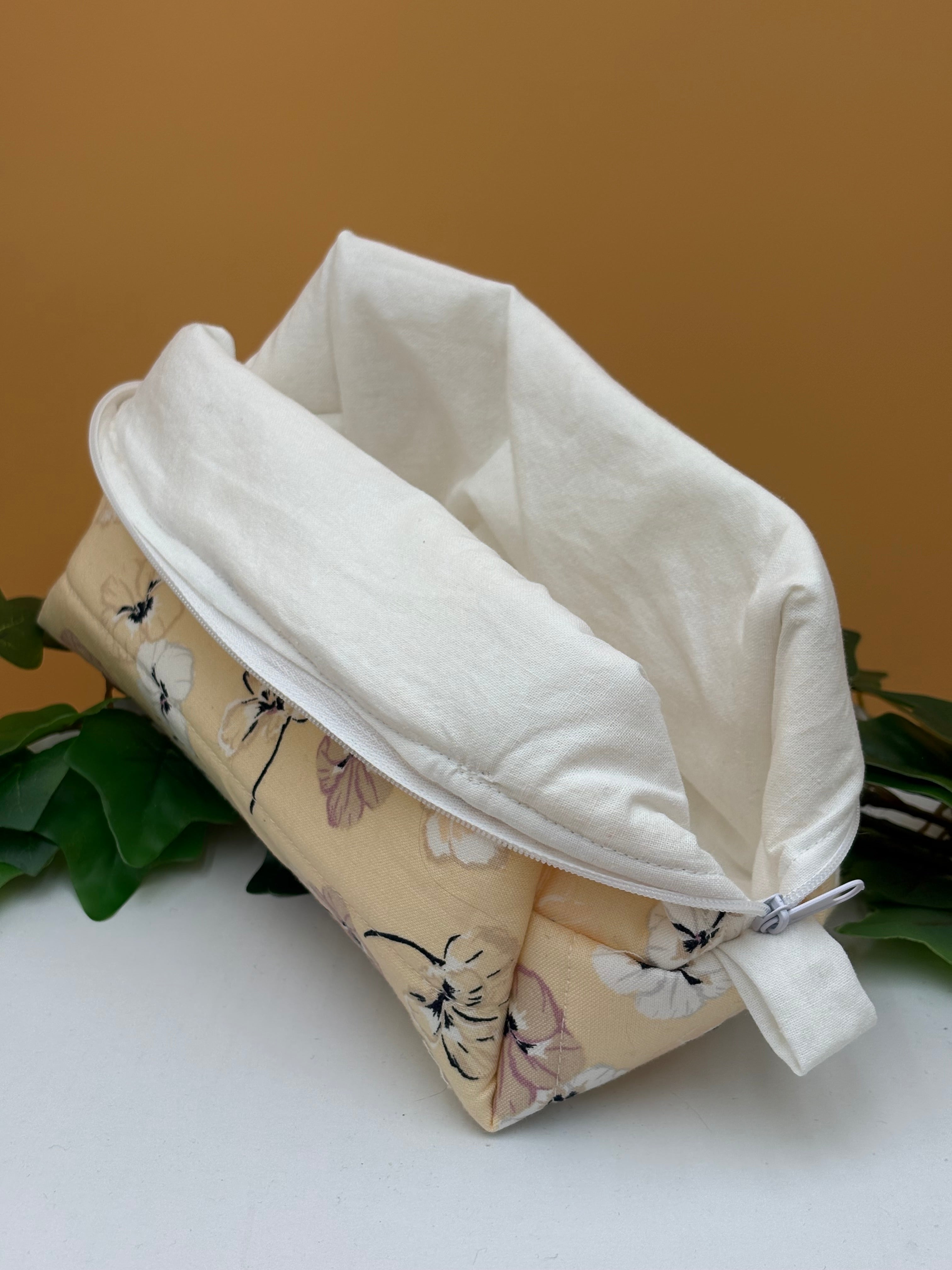 Quilted Pale Peach Floral pattern boxy makeup bag with white lining exposed on a tan background with a potted green plant