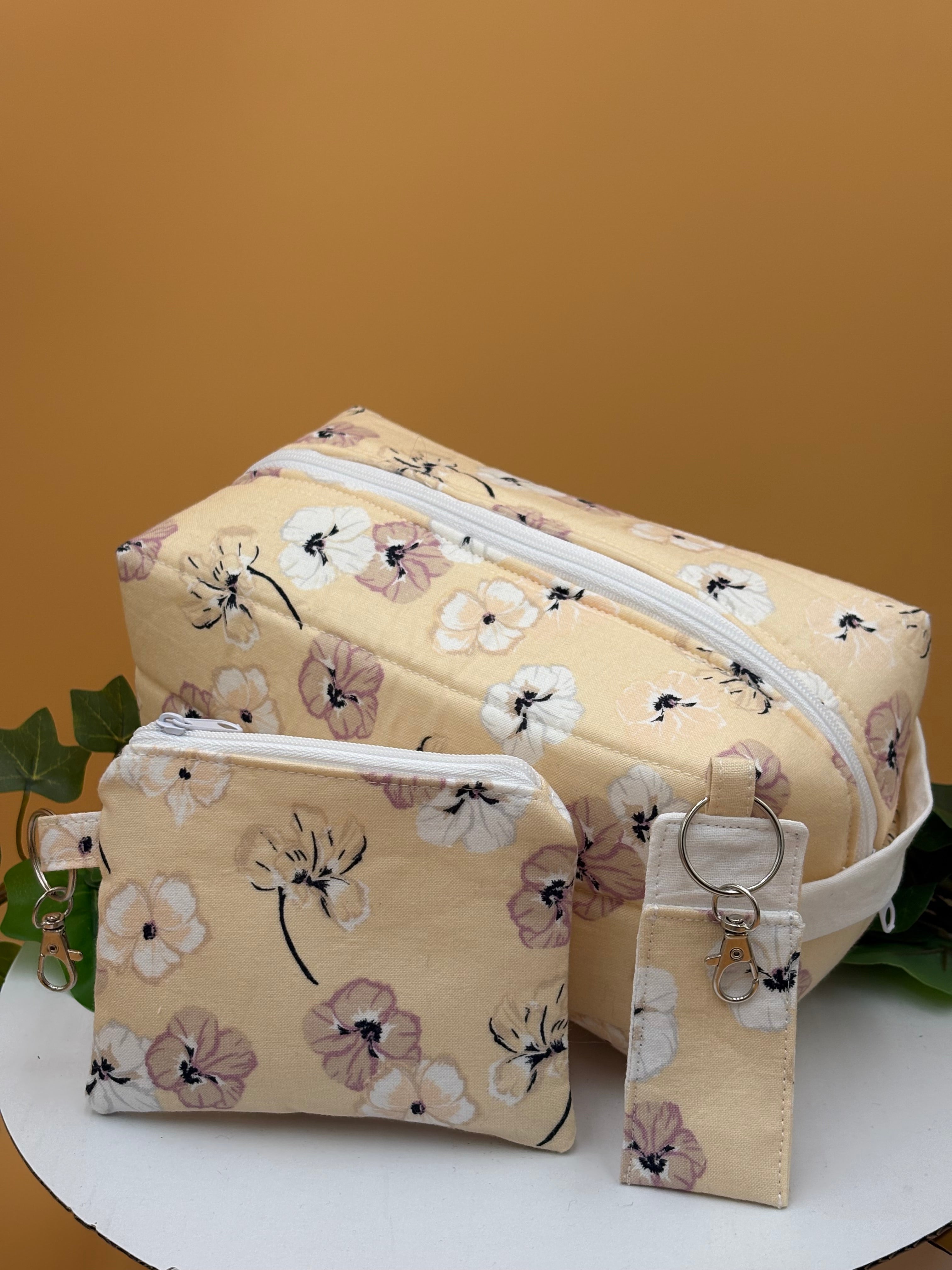 Quilted Pale Peach Floral pattern boxy makeup bag, coin purse keychain, and chapstick holder keychain with a on a tan background with a potted green plant