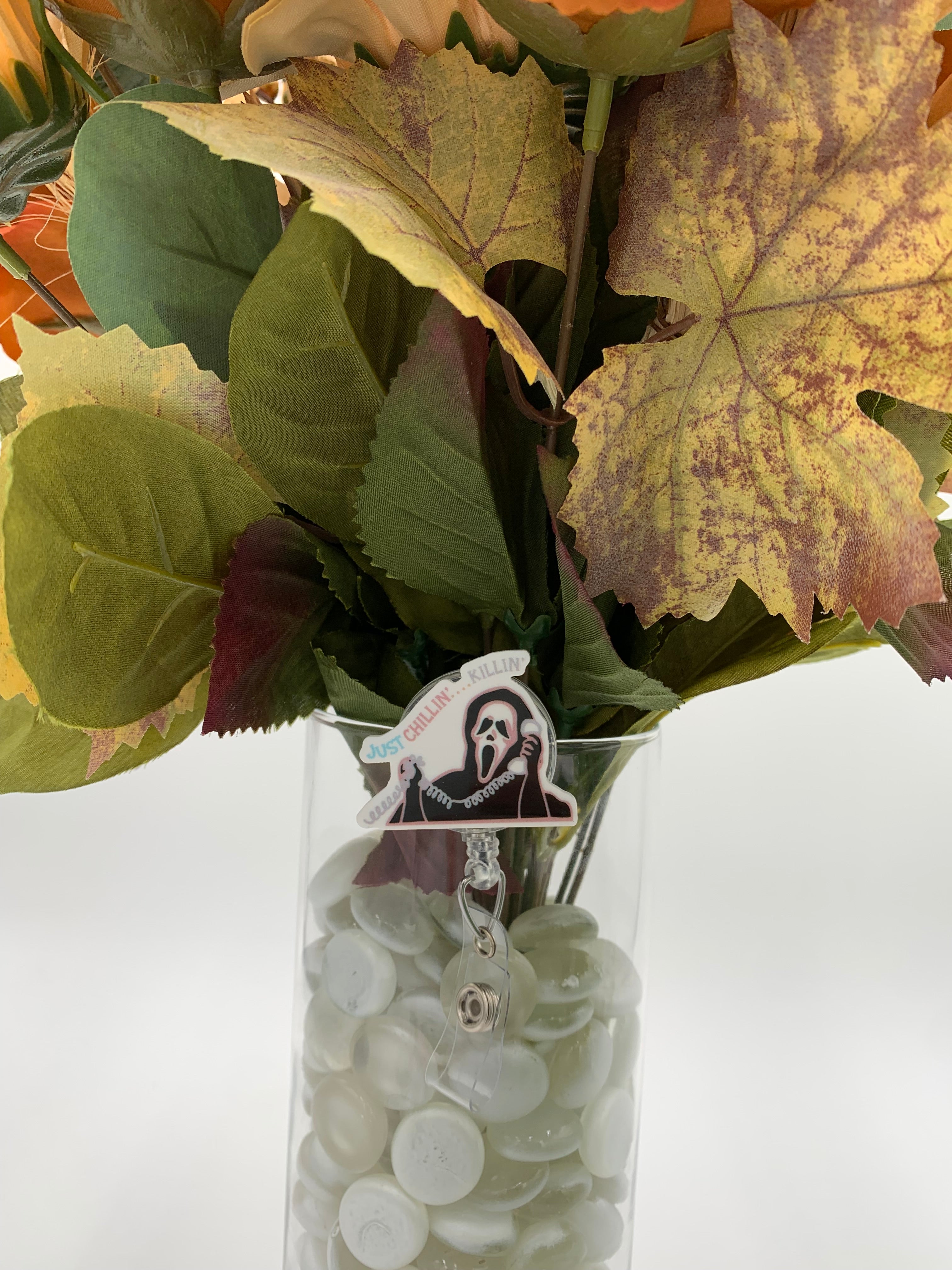 Bouquet of flowers with a man hooded man in a white mask holding a corded phone with text "just chillin'...killin'" badge holder in a clear vase on a white background