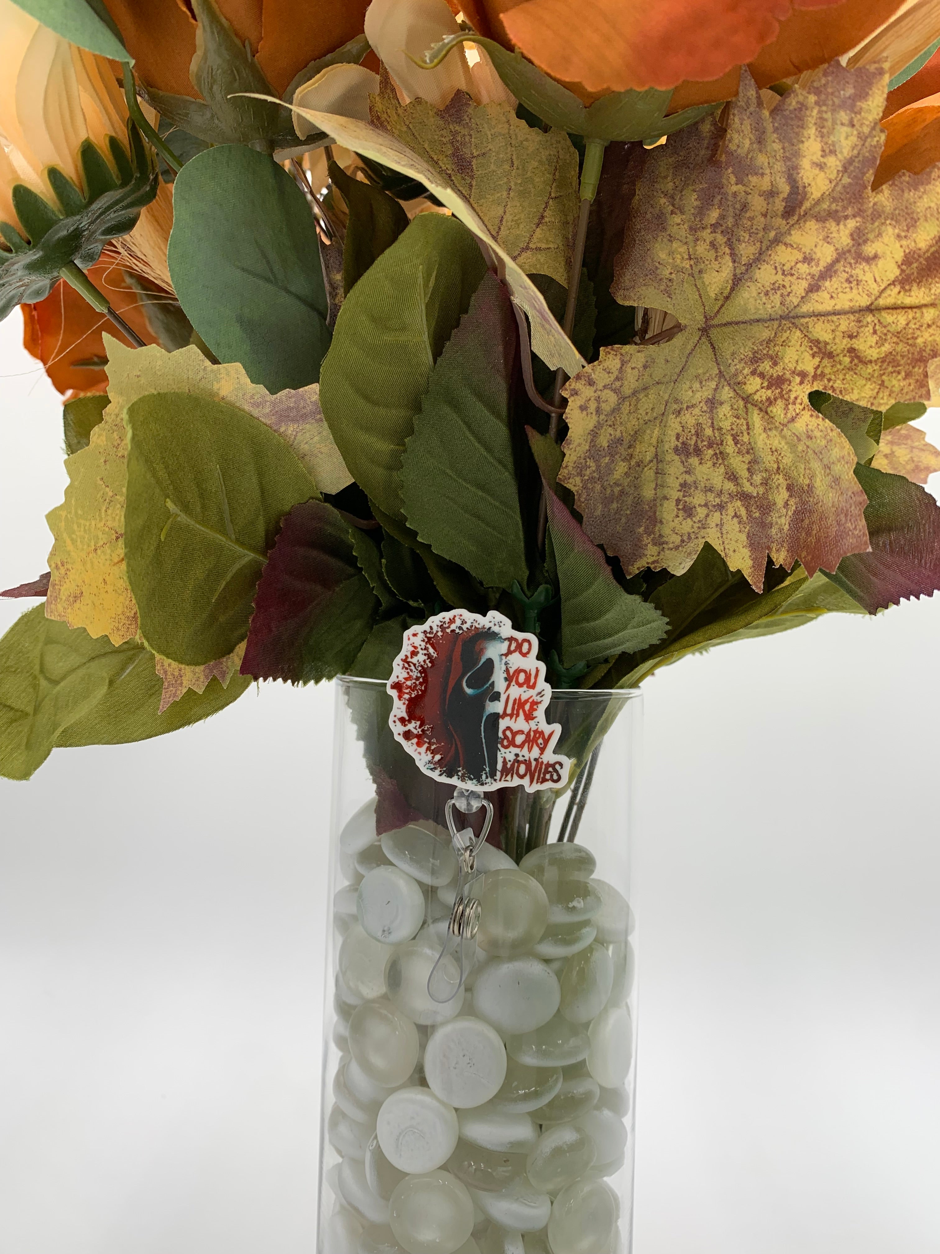 Bouquet of flowers with a hooded man in a white mask with text "do you like scary movies" badge holder in a clear vase on a white background