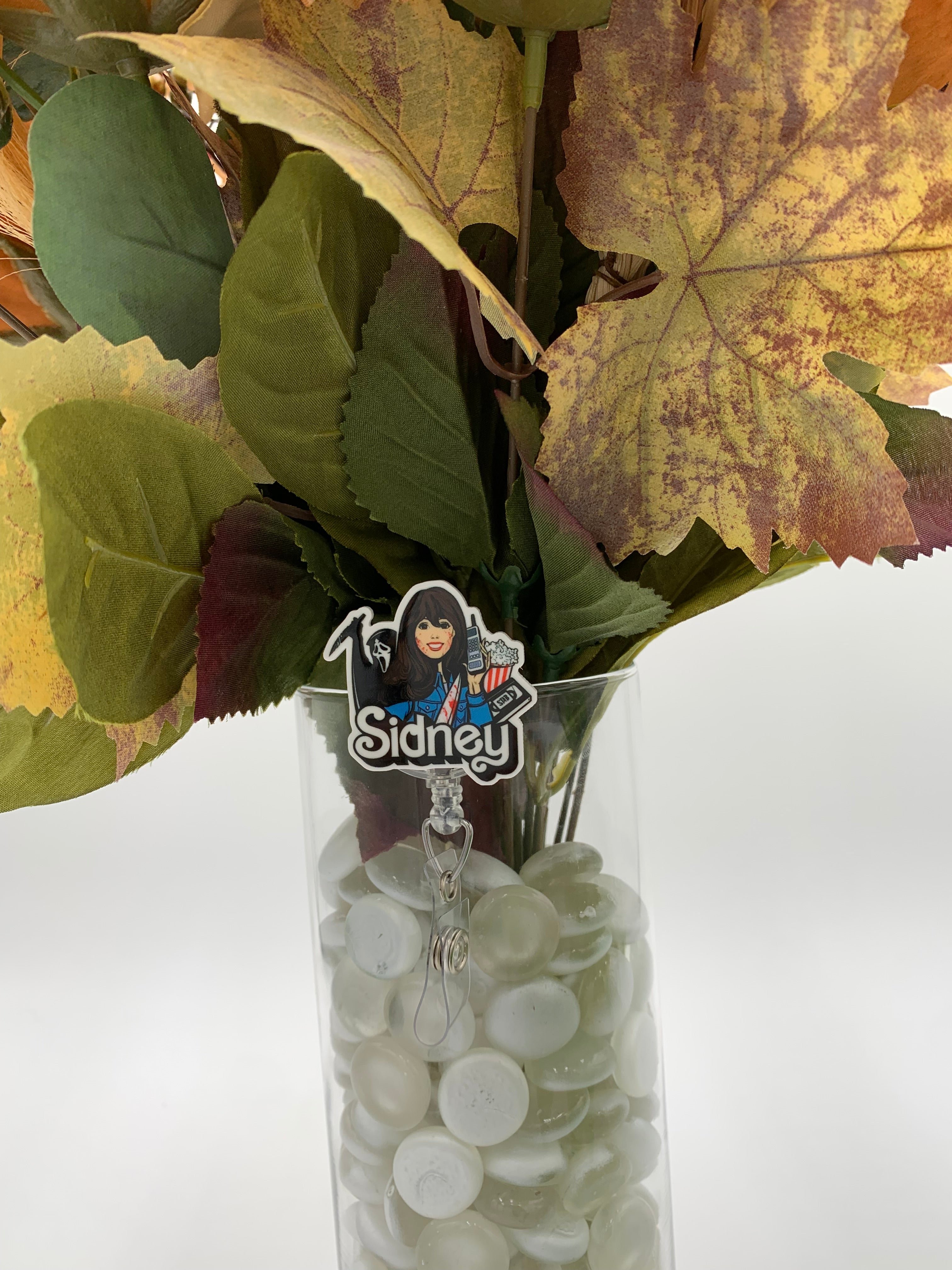 Bouquet of flowers with a hooded man with a white mask holding a knife next to a woman holding a cordless phone with a VHS and popcorn with text "Sidney" badge holder in a clear vase on a white background