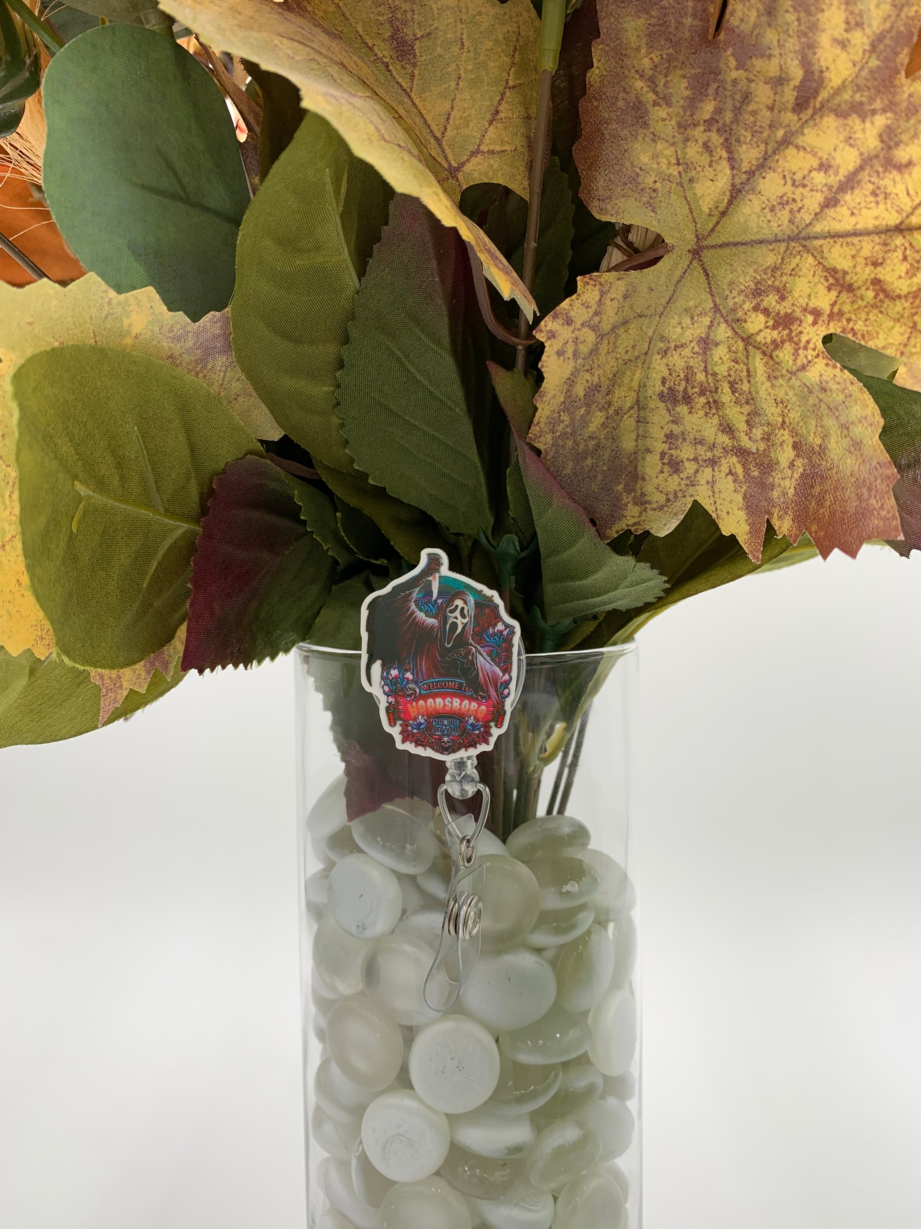 Bouquet of flowers with a hooded man with a white mask holding a knife with text "welcome to Woodsboro" badge holder in a clear vase on a white background