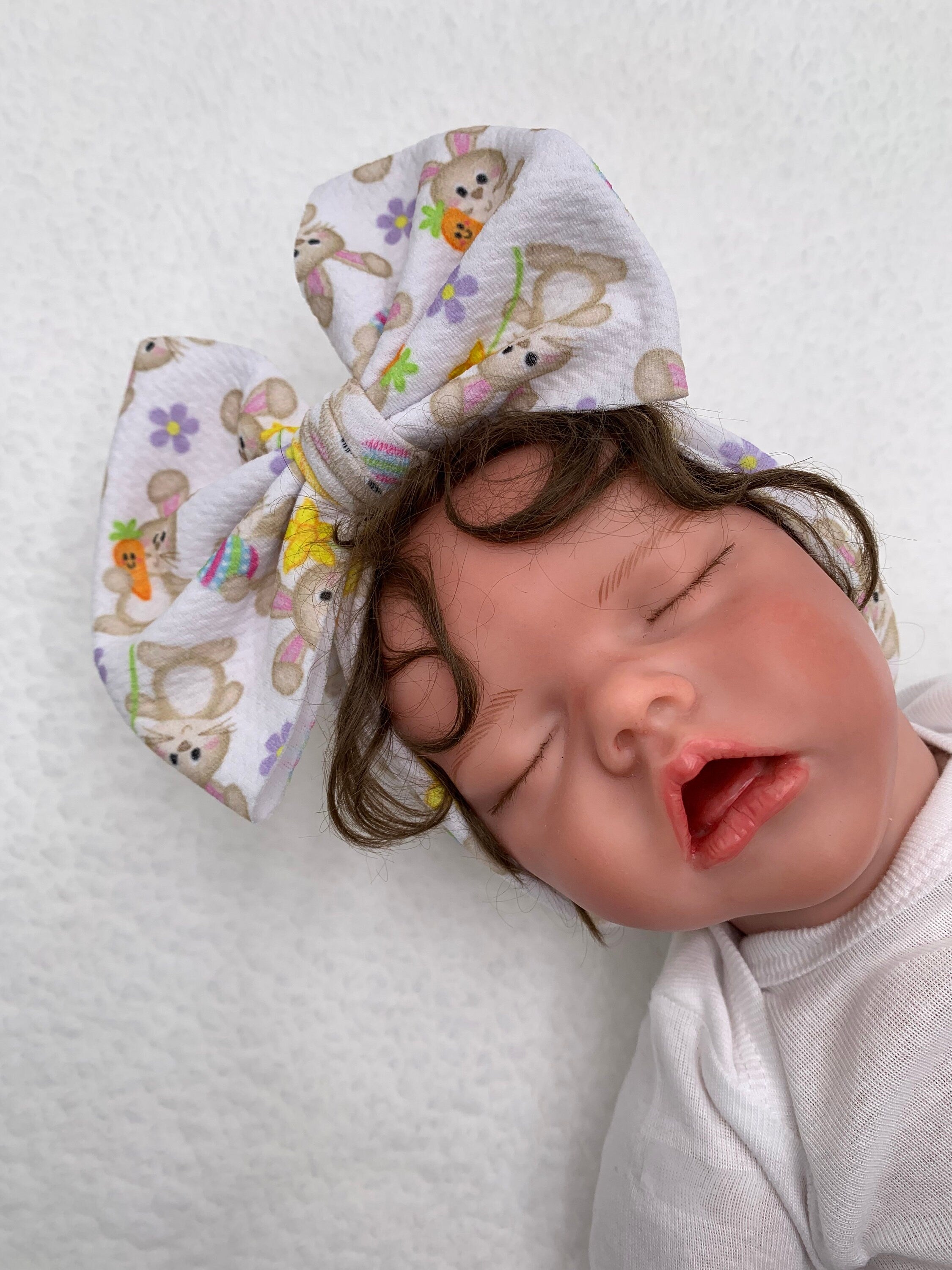 Newborn baby doll sleeping with a White Headband with a decorative bunny holding a carrot themed big bow on a mannequin head silhouette against a white background