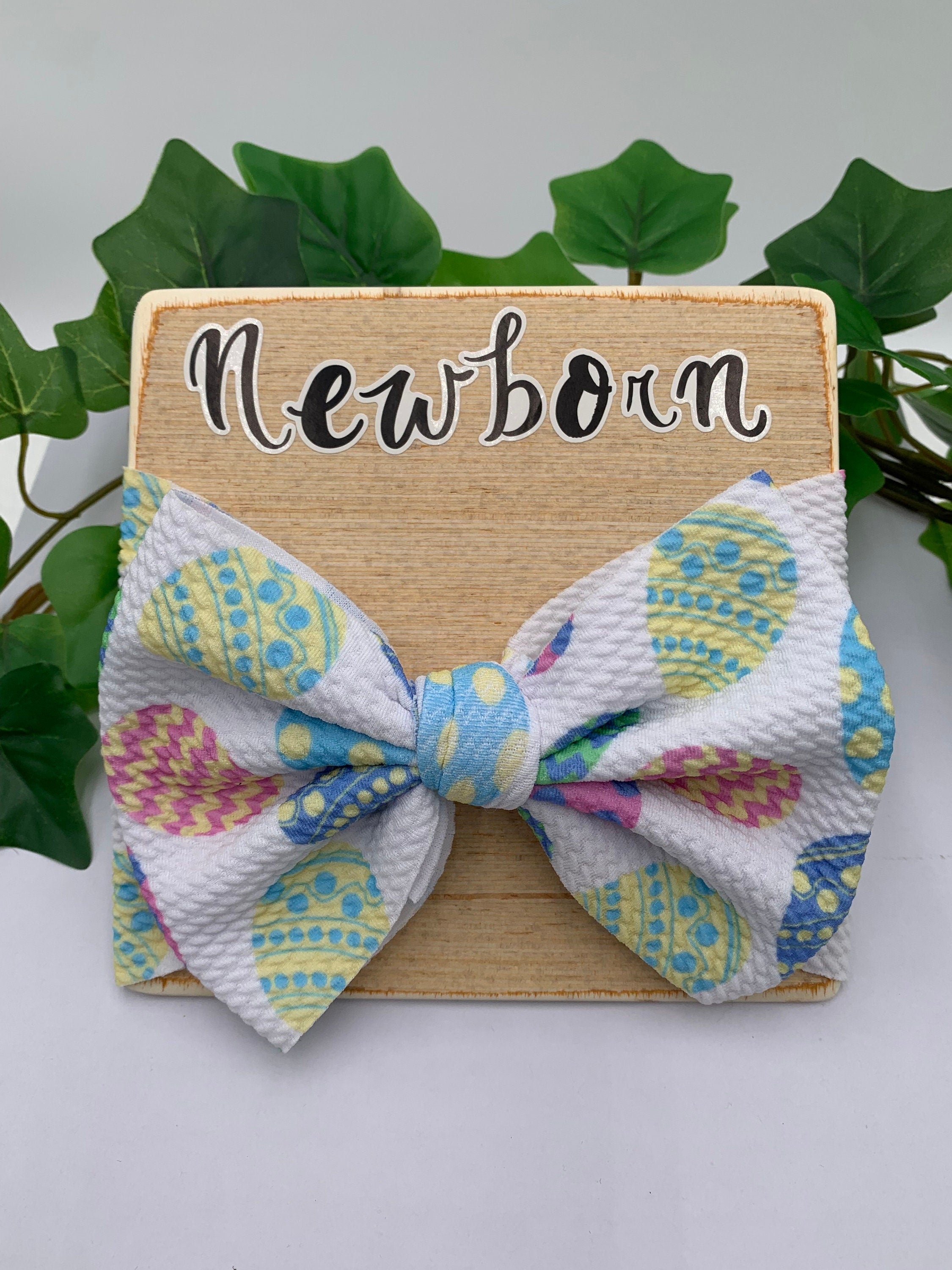 White Headband with a decorative easter eggs themed big bow on a wooden board with text "Newborn" against a white background