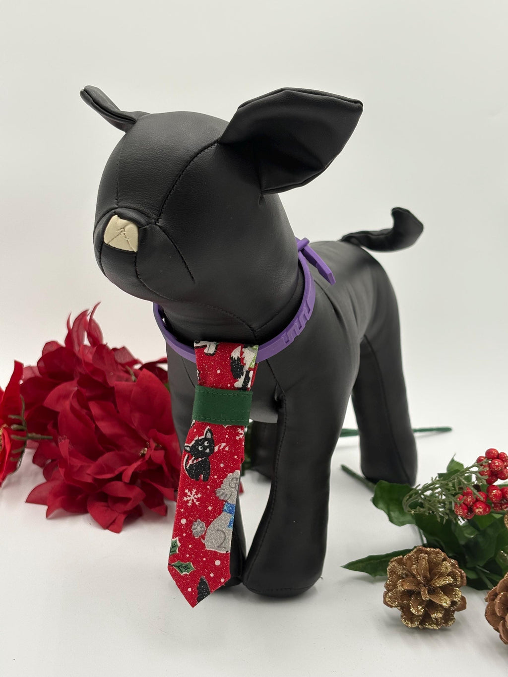 Black dog mannequin wearing a red over the collar necktie with a dog christmas pattern, surrounded by red flowers and greenery on a white background.