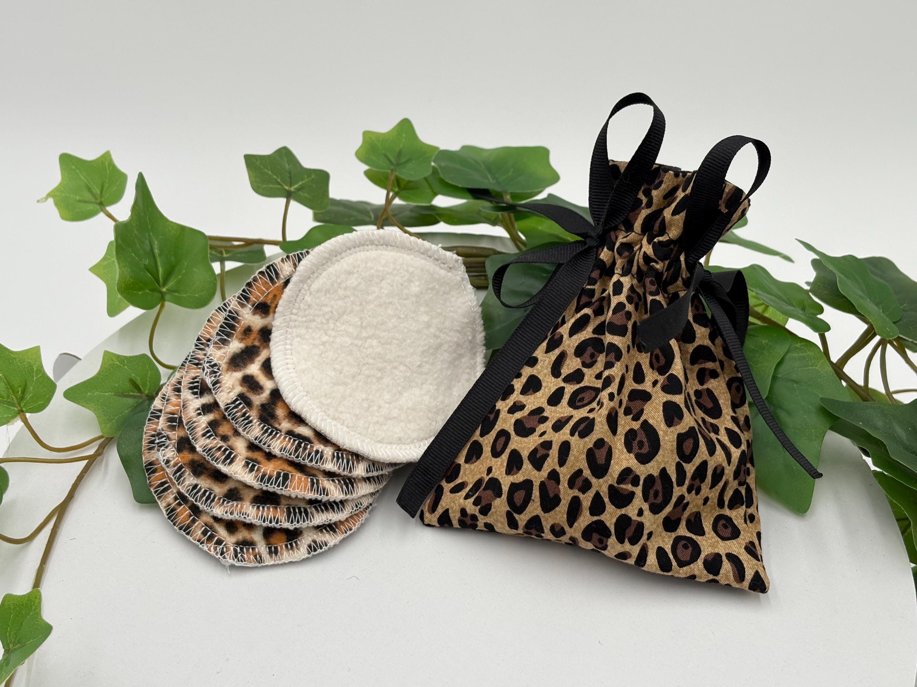Set of five reusable makeup remover pads with leopard print and white fabric with a leopard print drawstring bag, surrounded by green leaves on a white background.