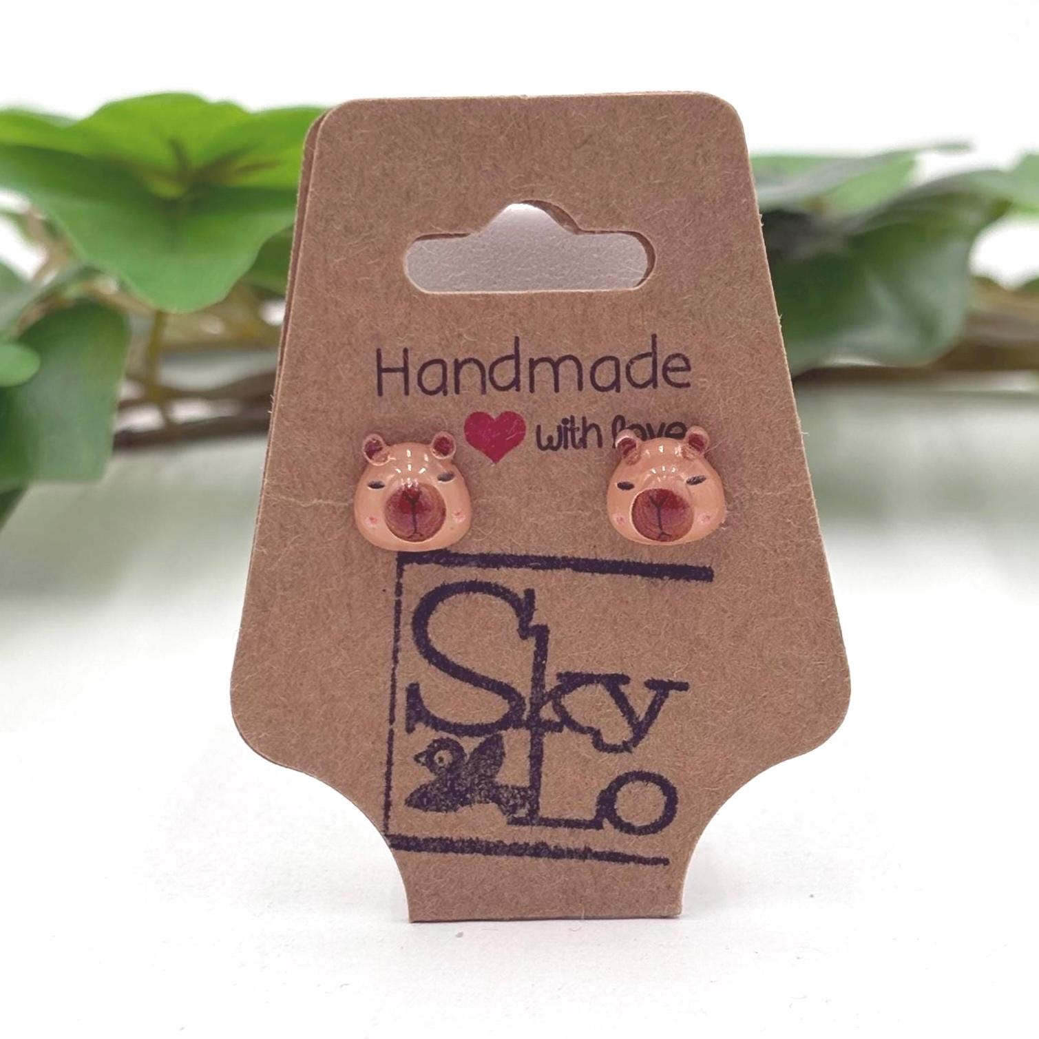 Handmade capybara head earrings on a branded card with a blurred green plant background