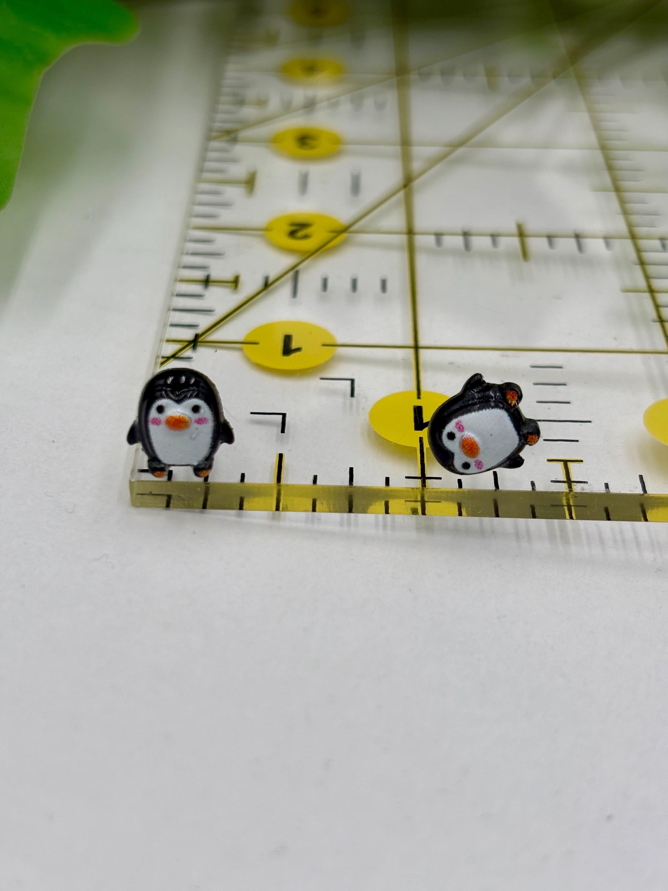 Two penguin earrings on a ruler for scale on a white background