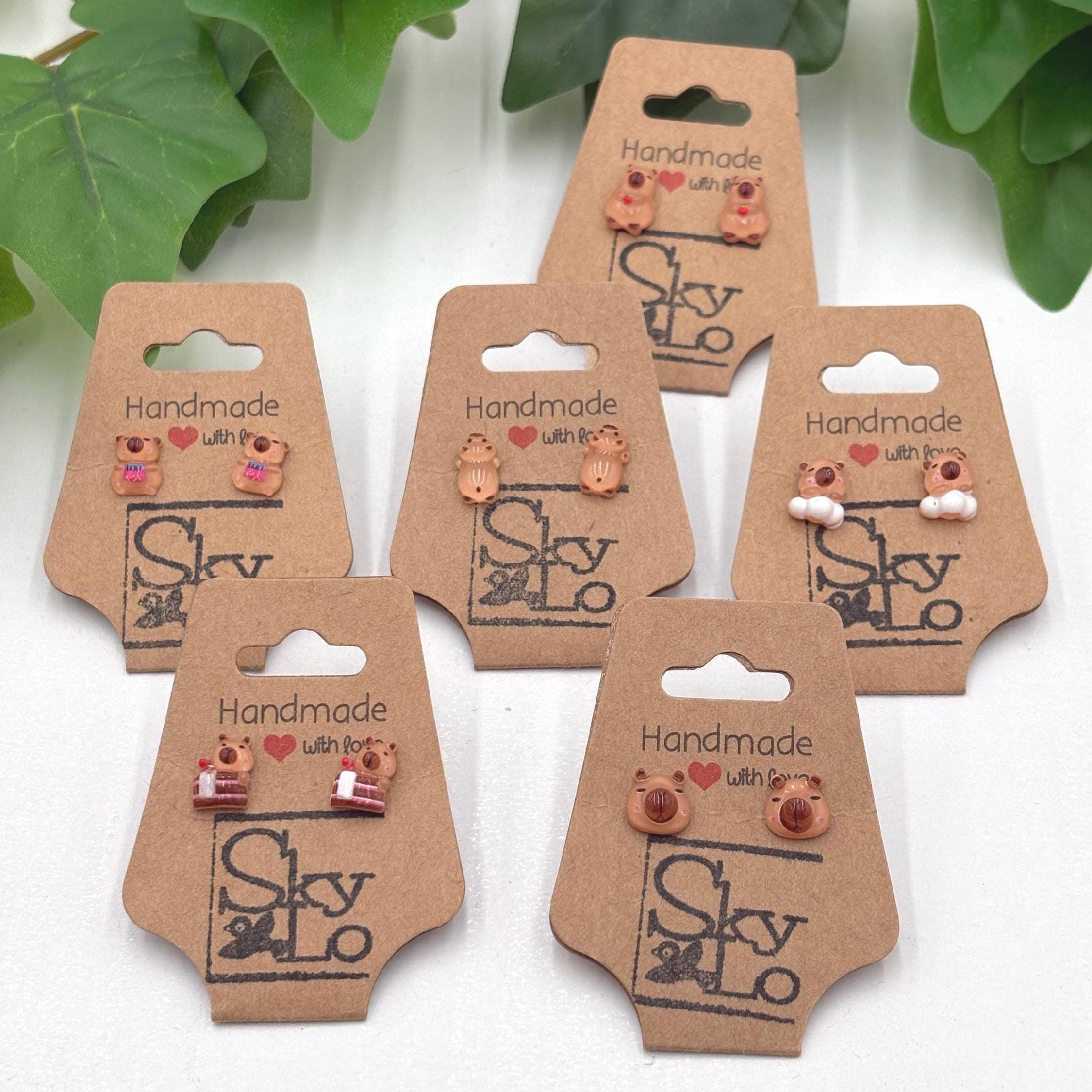 Set of six handmade capybara earrings on brown card holders with 'SkyLo' branding.
