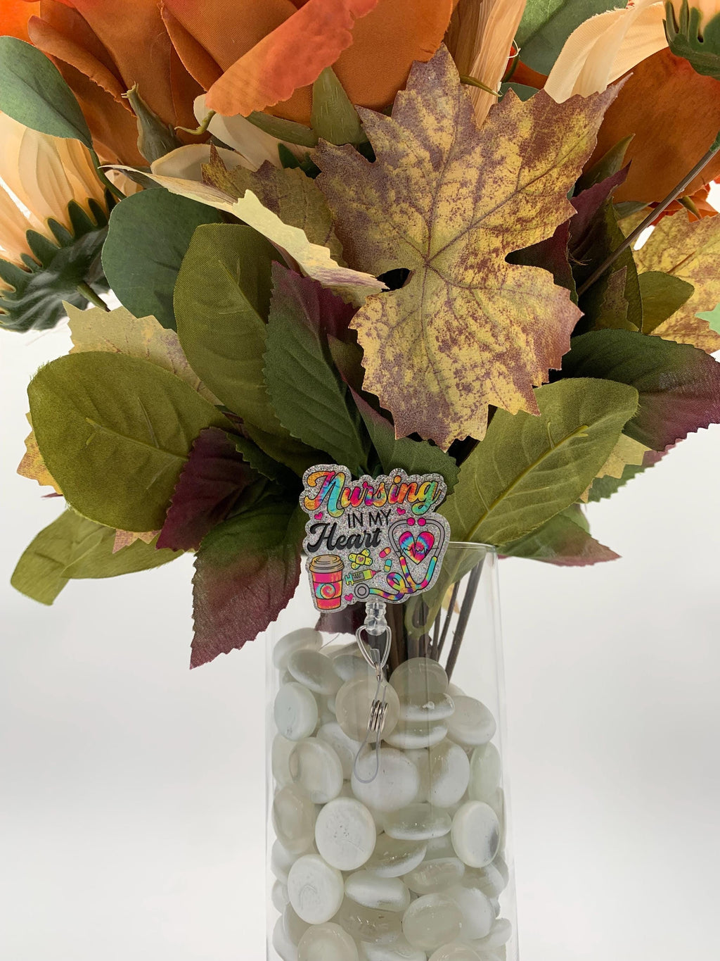Vase with autumn-themed flowers and a decorative charm on a white background with Badge reel that says "Nursing in my heart"