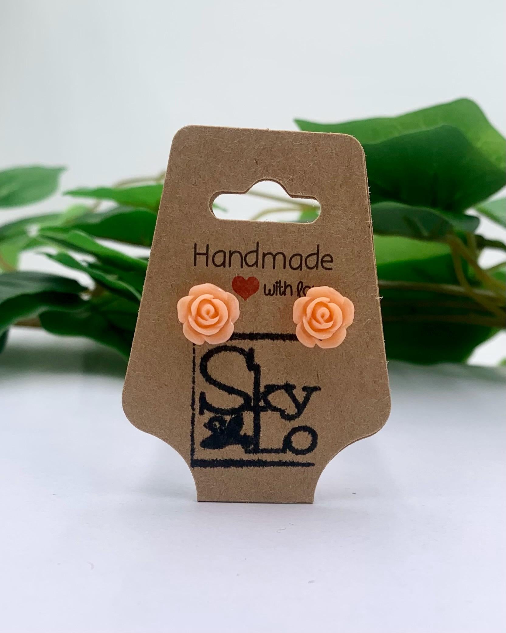 Pair of peach-colored rose-shaped earrings on a brown cardboard card with 'Handmade with Love SkyLo' branding, against a white background with green leaves.