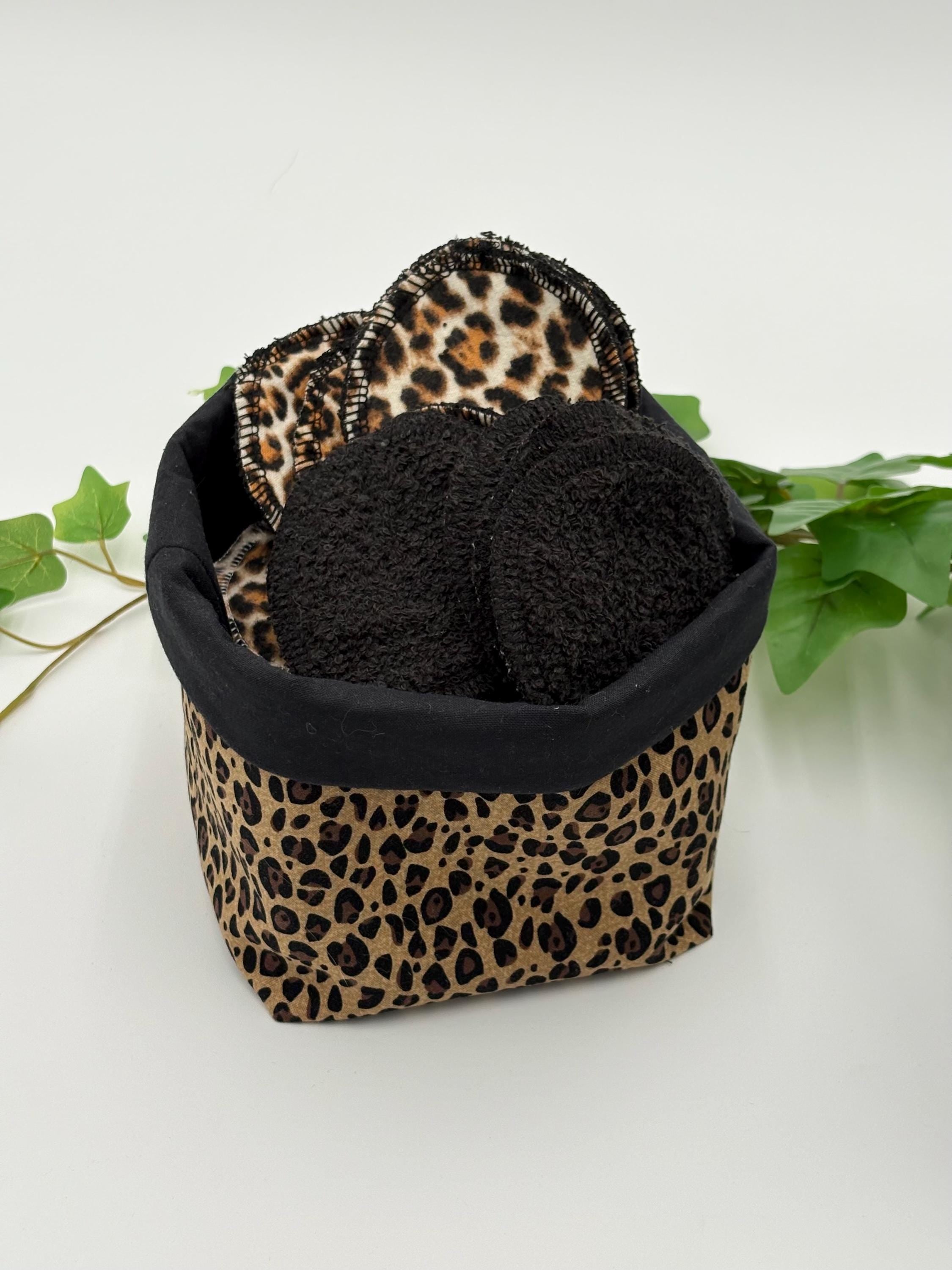 reusable makeup remover pads with leopard print and black fabric in a leopard print and black fabric basket, surrounded by green leaves on a white background.