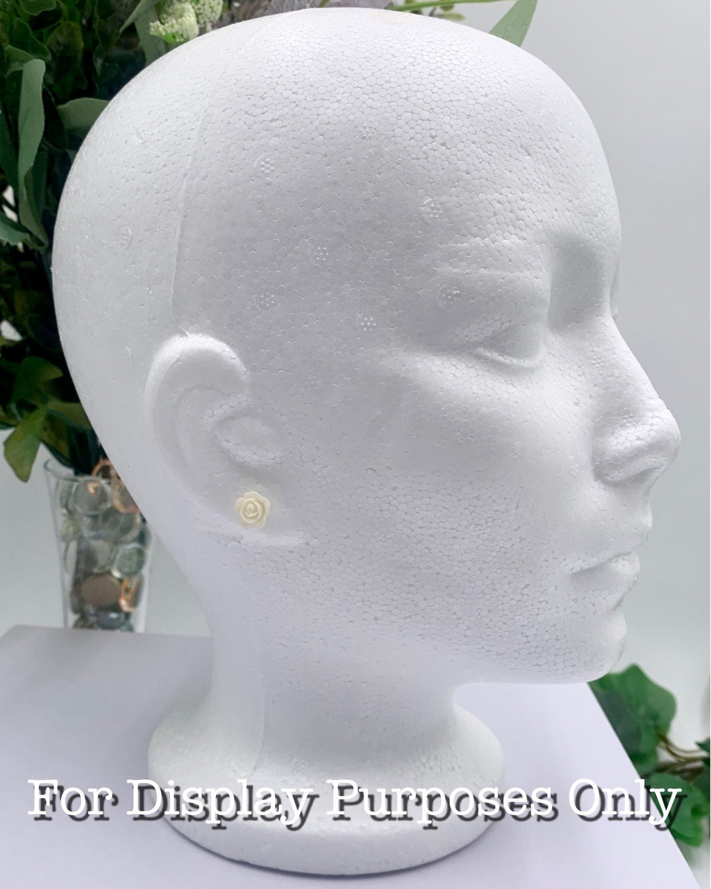White mannequin head with a small butter cream rose earring on a white background with greenery with text "for display purposes only"