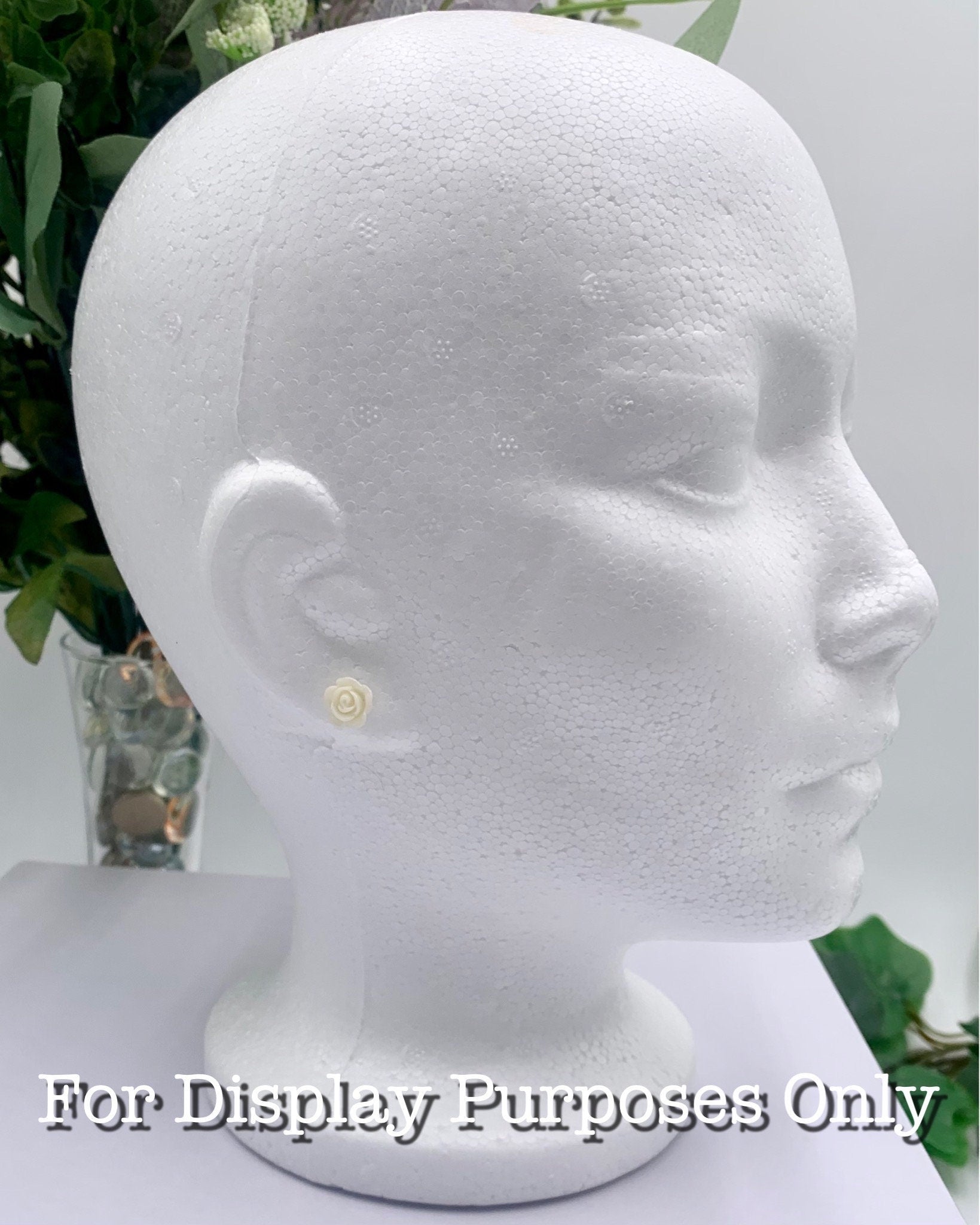 White mannequin head with a small butter cream rose earring on a white background with greenery with text "for display purposes only"