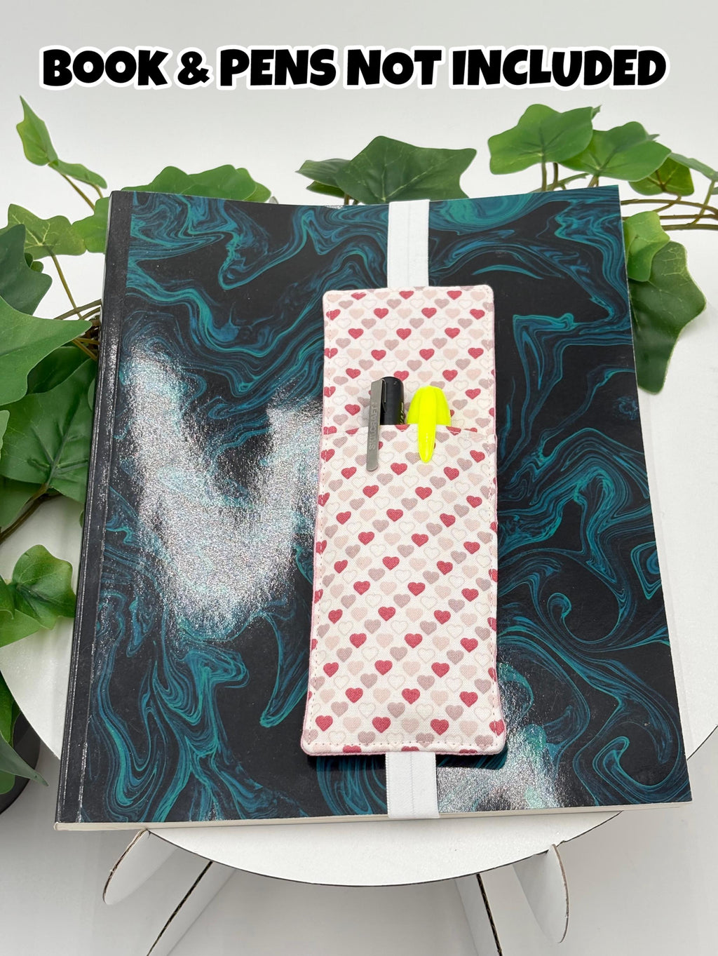 Pink pen holder book mark with hearts pattern on a marbled black and blue surface, with text 'Book & Pens Not Included'.