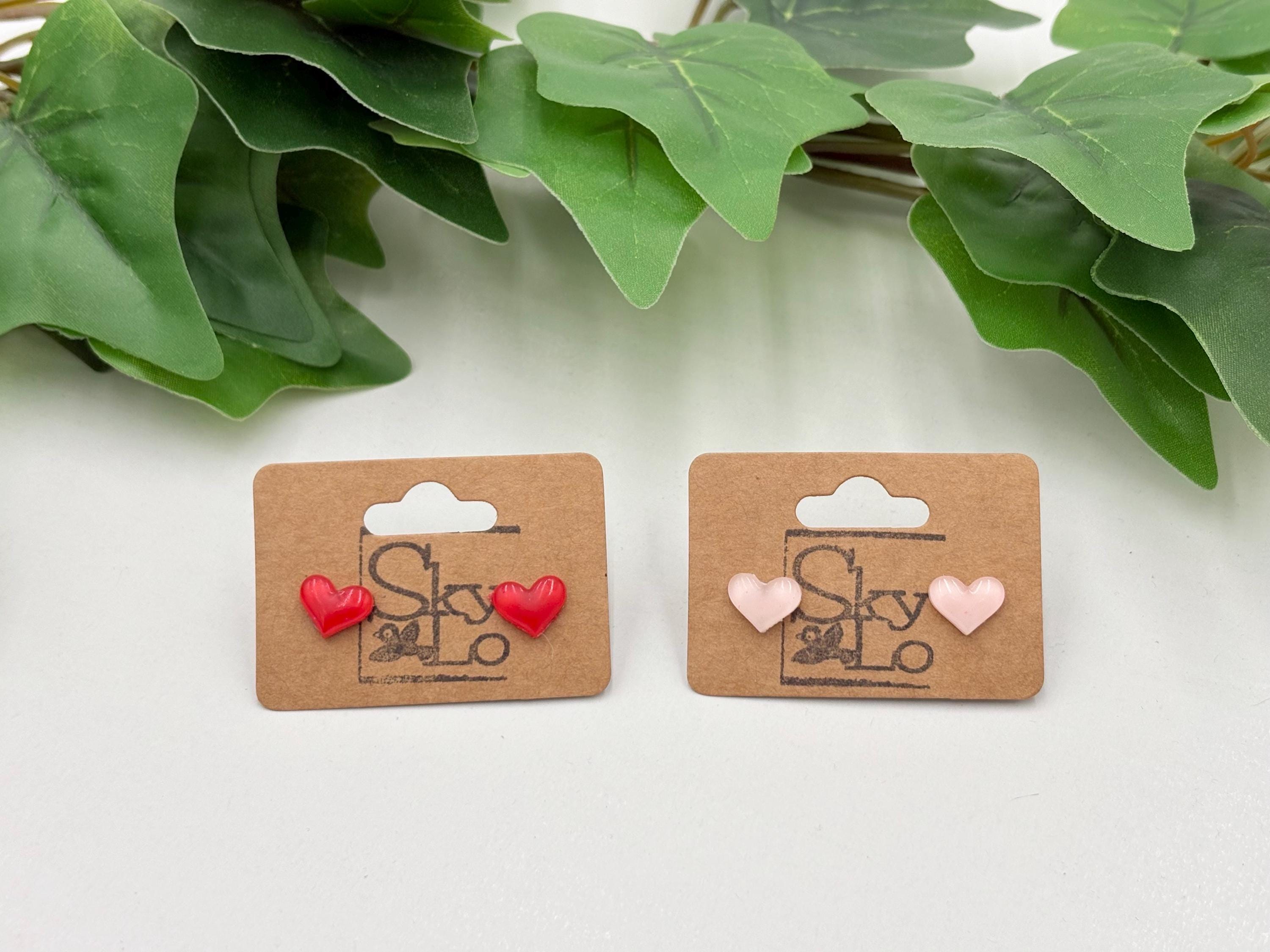 Two pairs of heart-shaped earrings on branded cards with green leaves in the background.