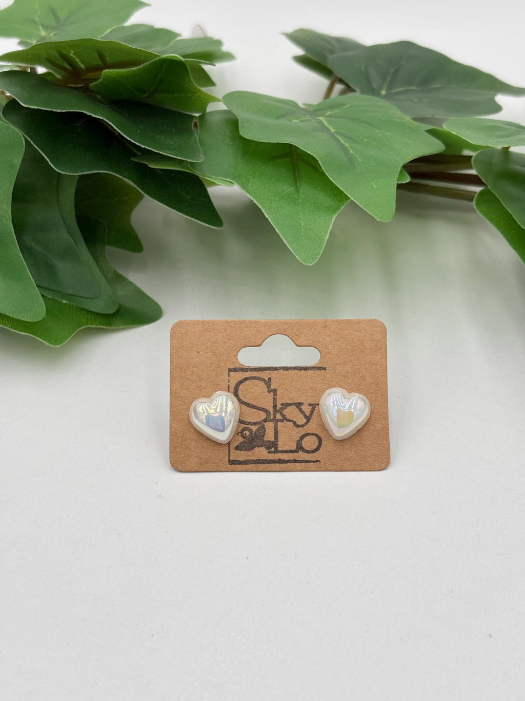 White metallic Heart-shaped earrings on display with a brand label against a white background with green leaves.