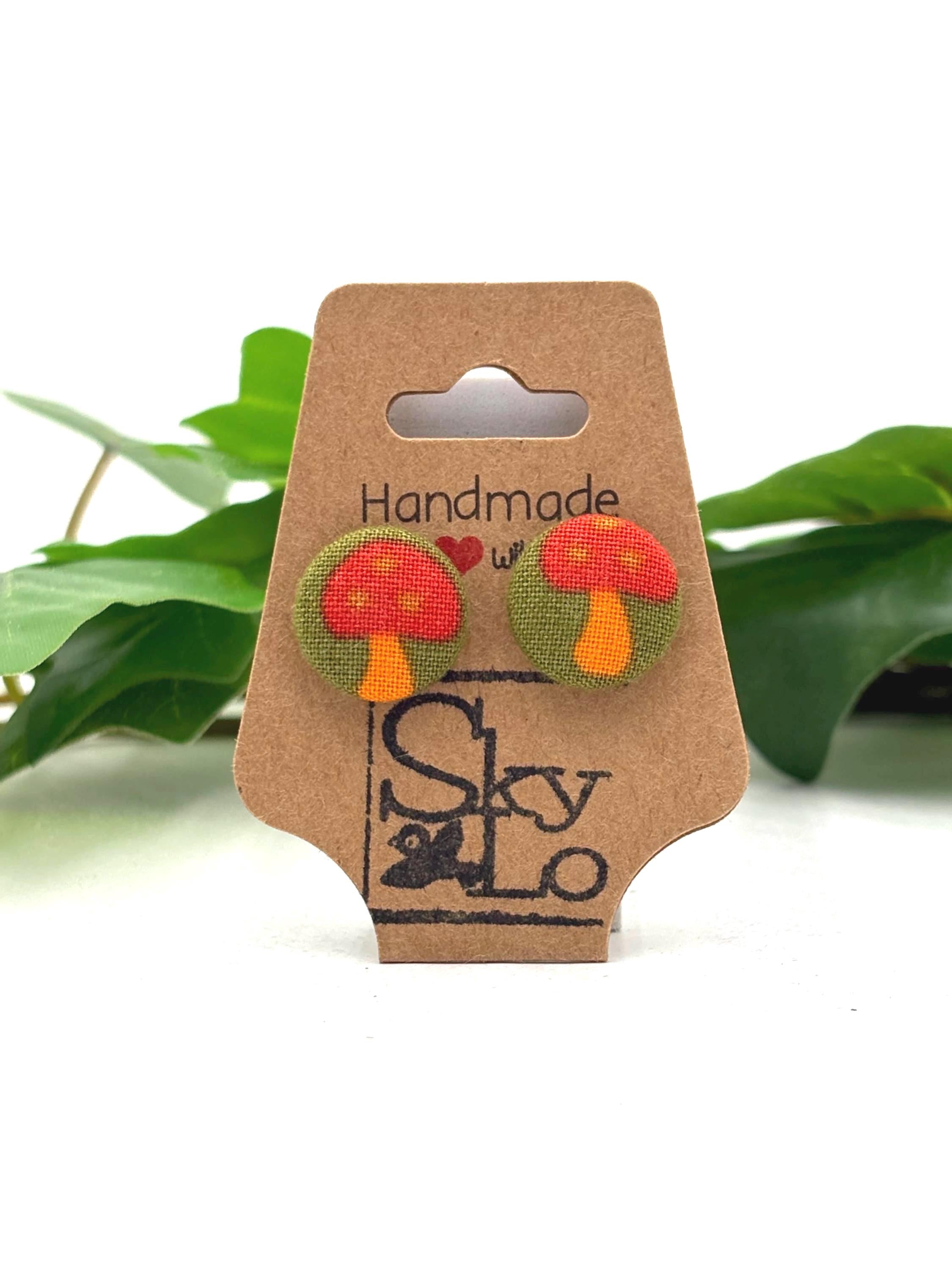 Cardboard earring display card with red mushroom earrings and 'Handmade with love' and 'SkyLo' branding, against a white background with green leaves.