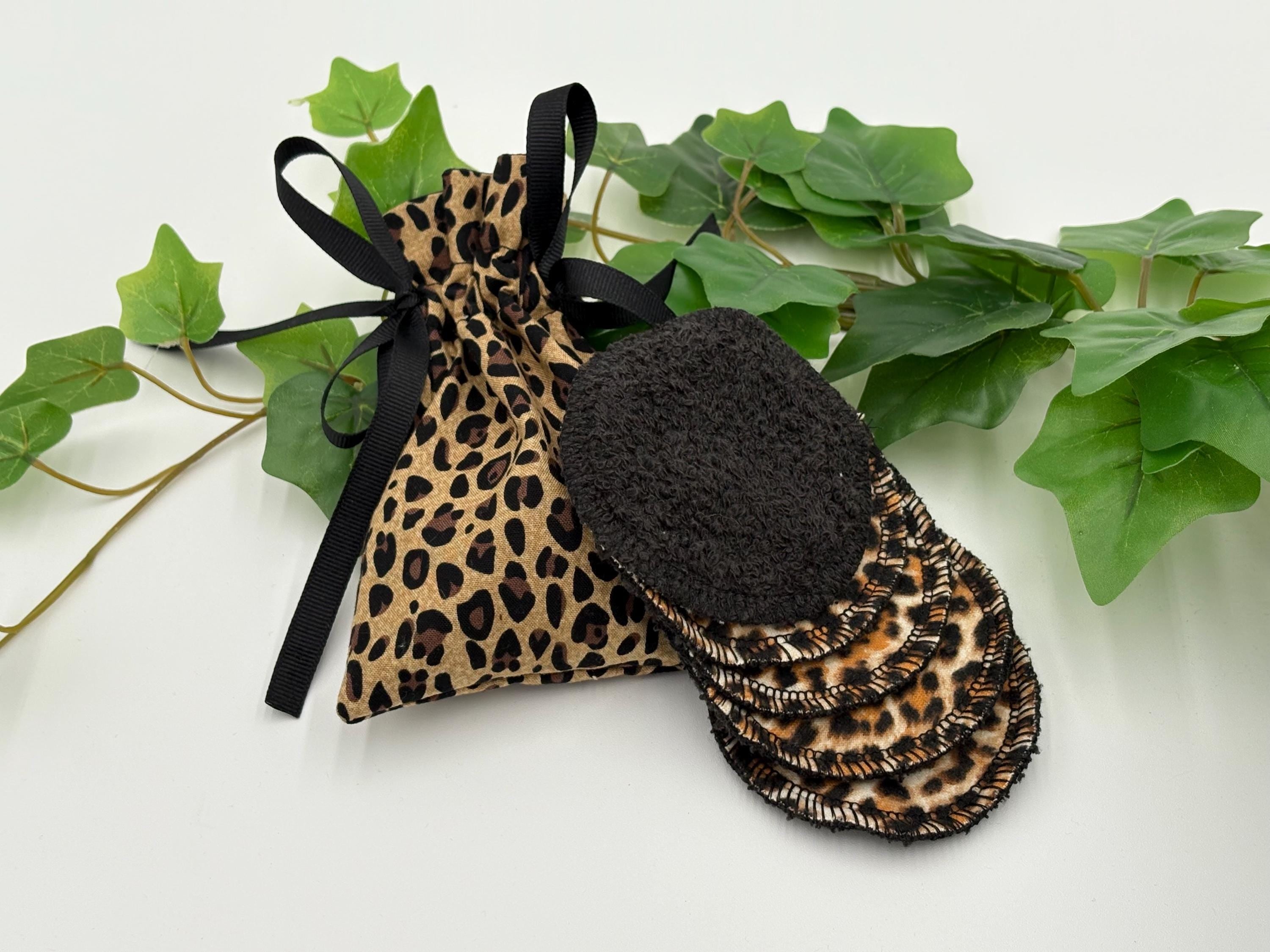Set of five reusable makeup remover pads with leopard print and black fabric with a leopard print drawstring bag, surrounded by green leaves on a white background.