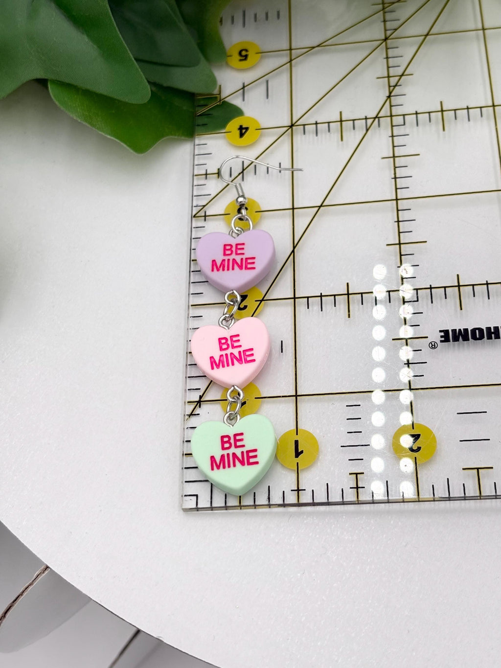 Heart-shaped dangle earring with text 'Be Mine' on a ruler with green leaves