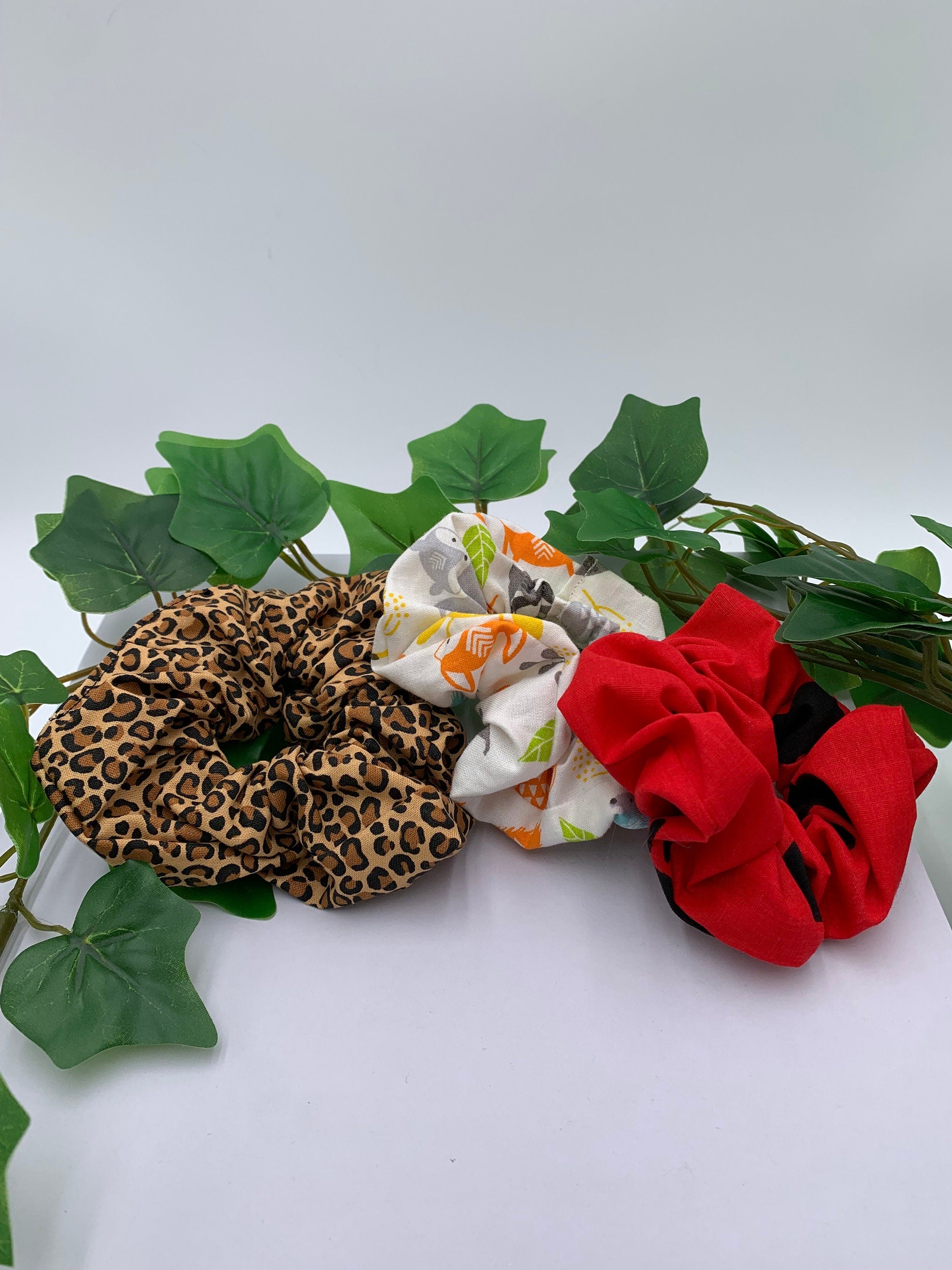 Set of three scrunchies with leopard print, white patterned woodland critters, and red with black paw prints designs on a white background next to green leaves